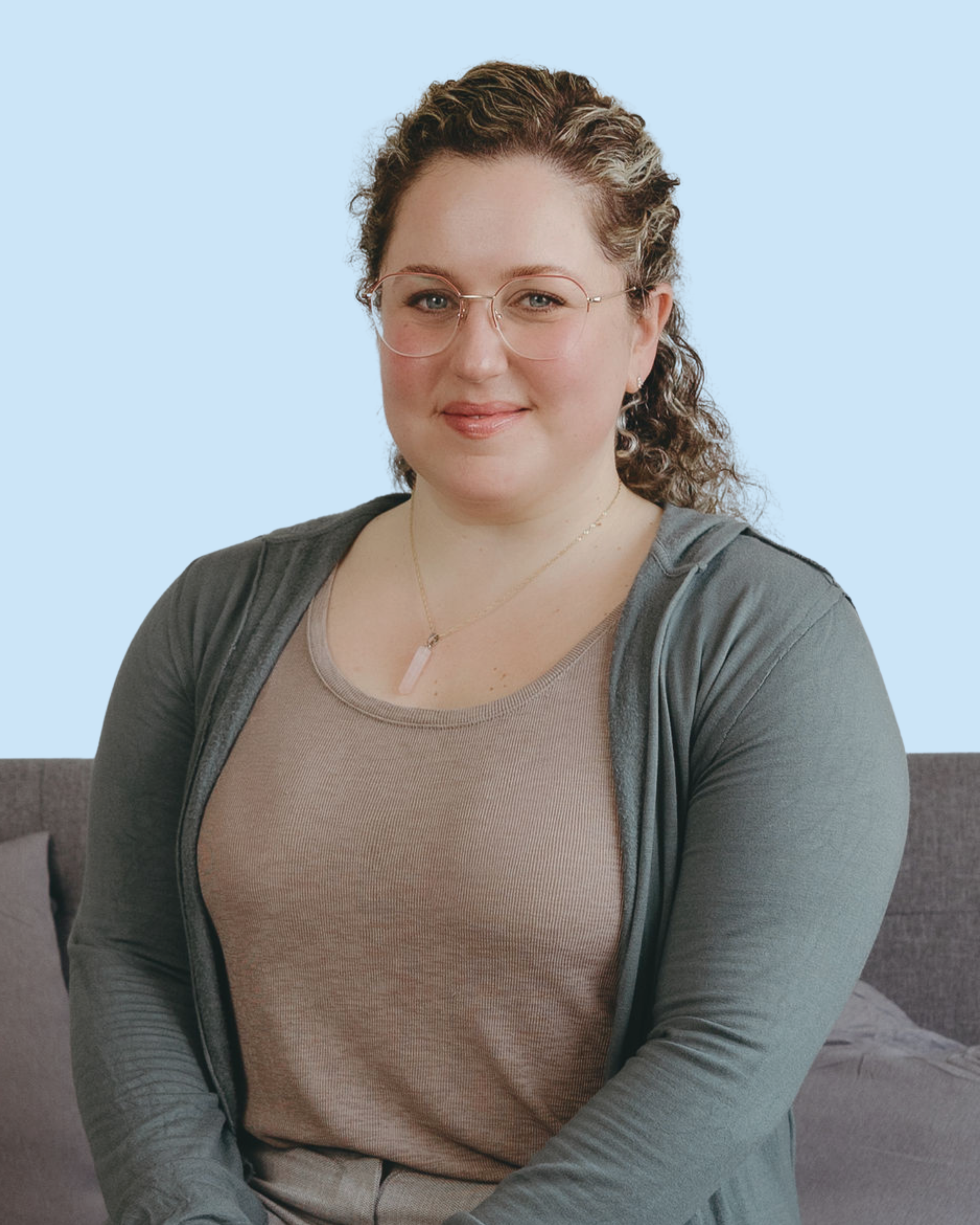 A woman with curly hair, glasses, and a gold necklace sitting on a gray couch against a light blue background.
