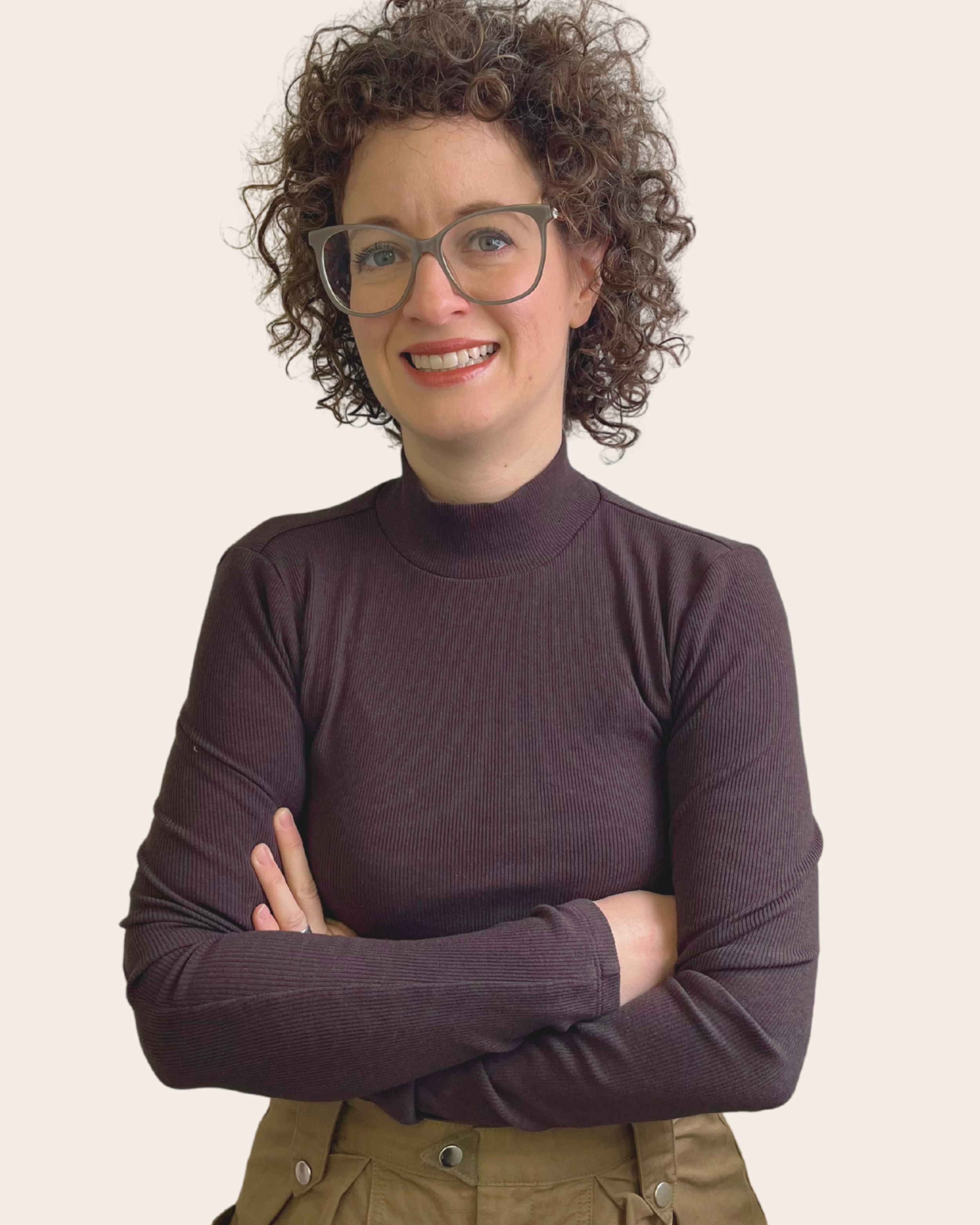 A woman with curly hair and glasses wearing a dark long-sleeve turtleneck, standing with arms crossed and smiling in front of a plain light background.