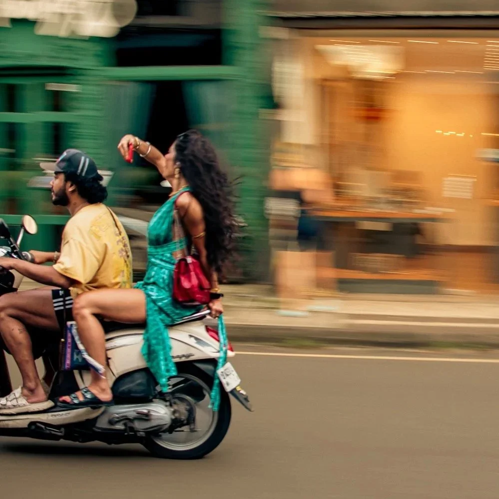 A man and woman riding a scooter on a city street, with the woman wearing a turquoise dress and the man wearing a yellow shirt and cap, against a blurred city background
