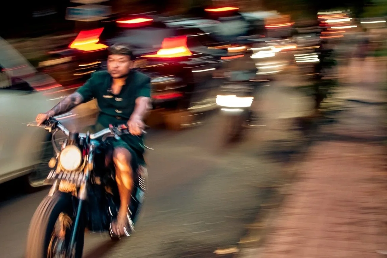 A man riding a motorcycle on a busy street at dusk, with cars and taillights creating blurred streaks of red and white lights in the background.