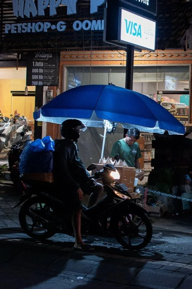Nighttime street scene with food stall illuminated by a light, two umbrellas (blue and red) shading the stall, motorcyclist wearing a helmet and carrying a backpack passing by, several parked scooters and motorcycles, a shop signage with a VISA sign,