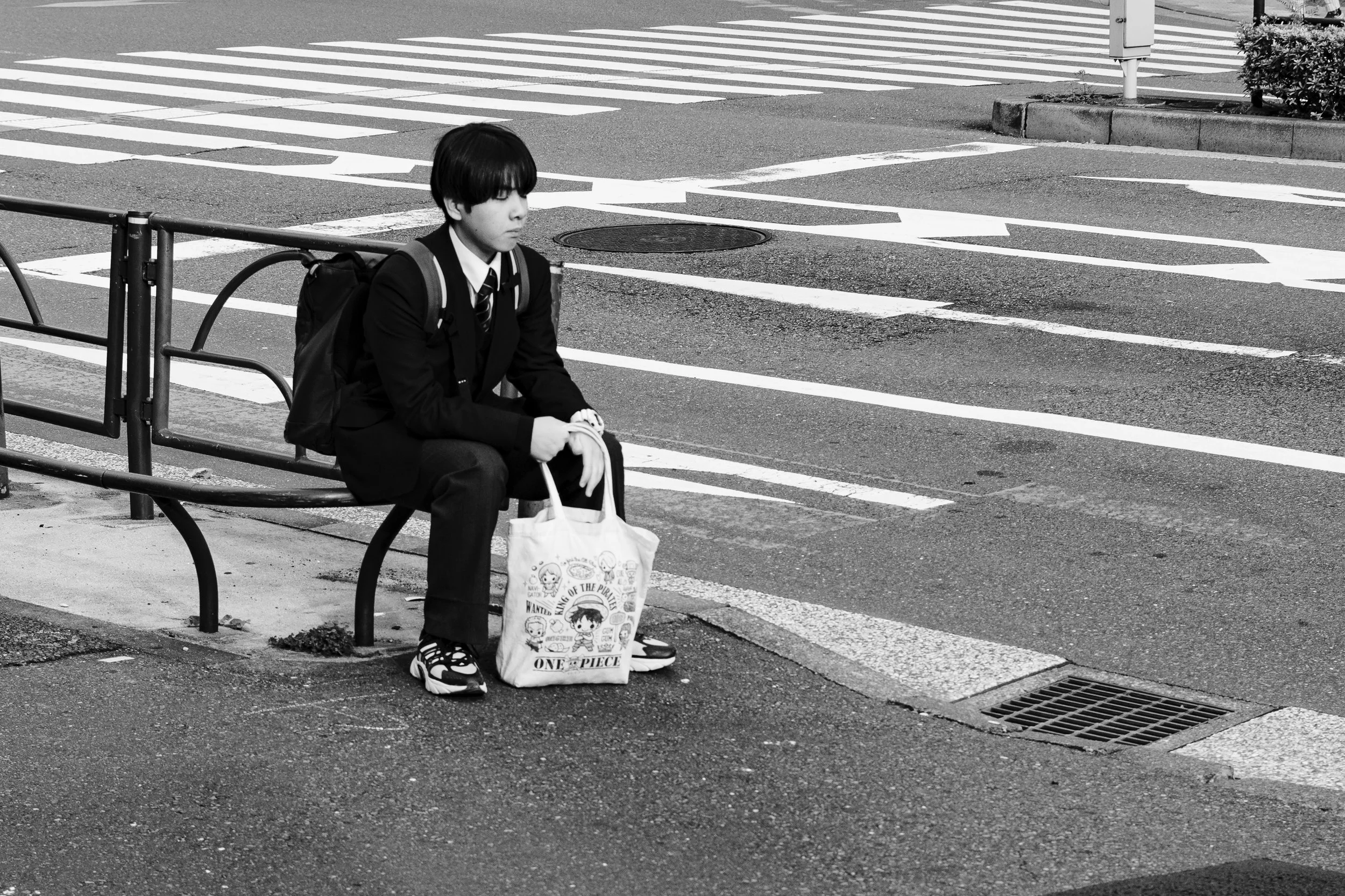 A young person in a school uniform sitting on a bench at a crosswalk, holding a tote bag with cartoon characters.