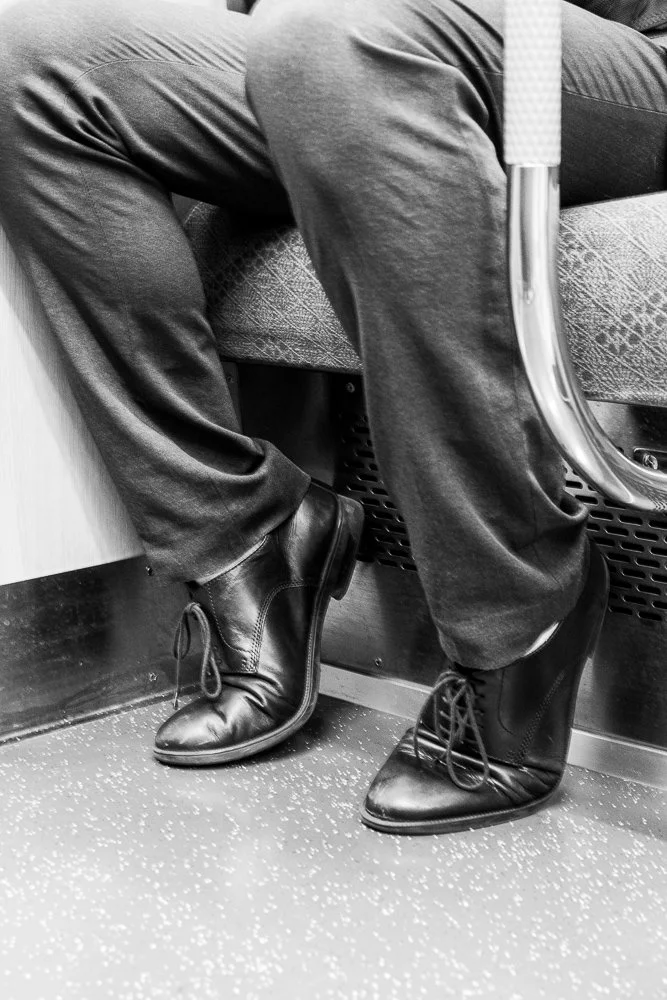 A person sitting on a train with legs crossed, wearing dark dress shoes and dark pants.