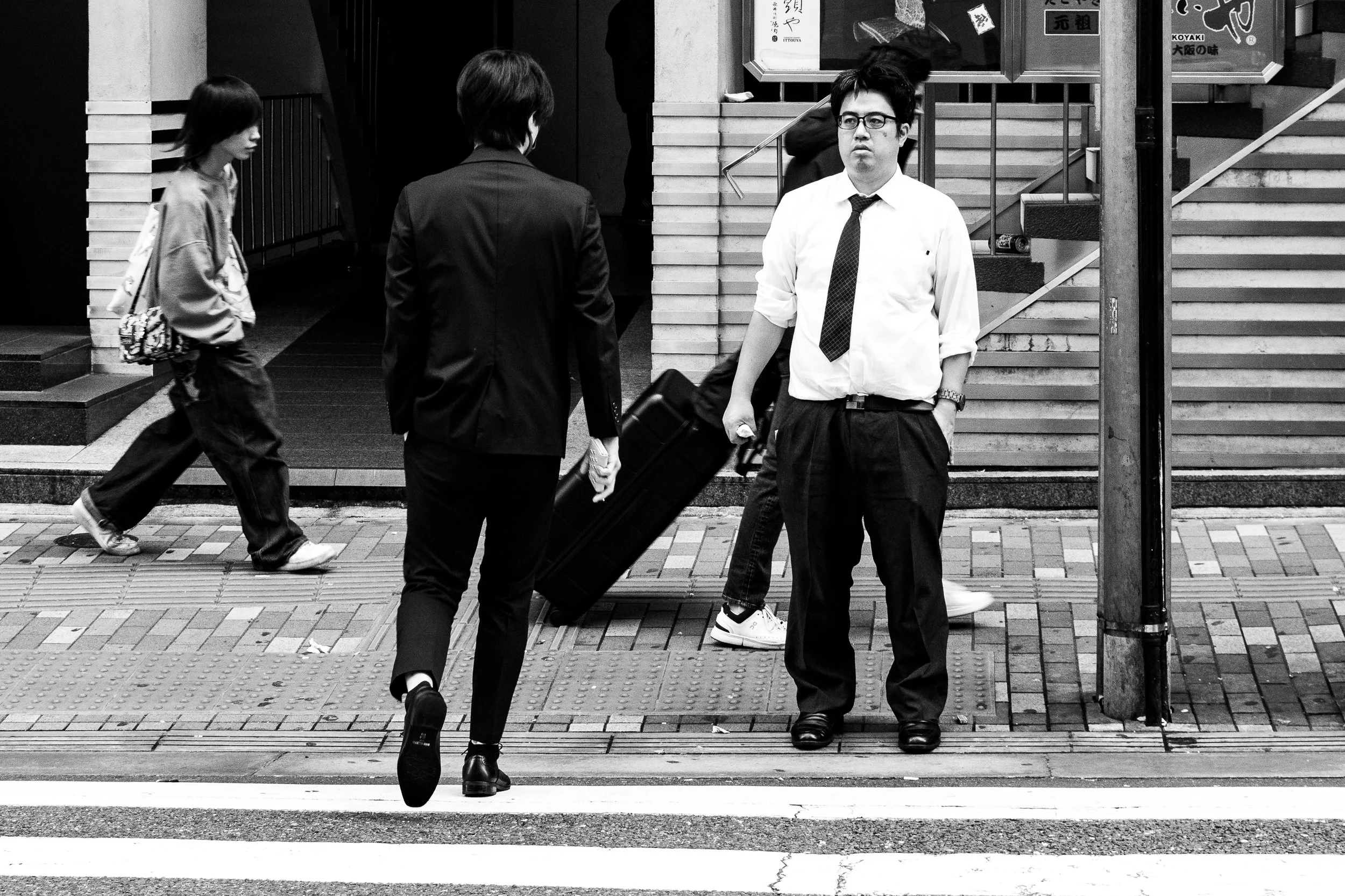 Black and white photo of people waiting at a crosswalk, including a man in a white shirt, tie, and glasses, and a woman in a dark suit pulling a rolling suitcase.