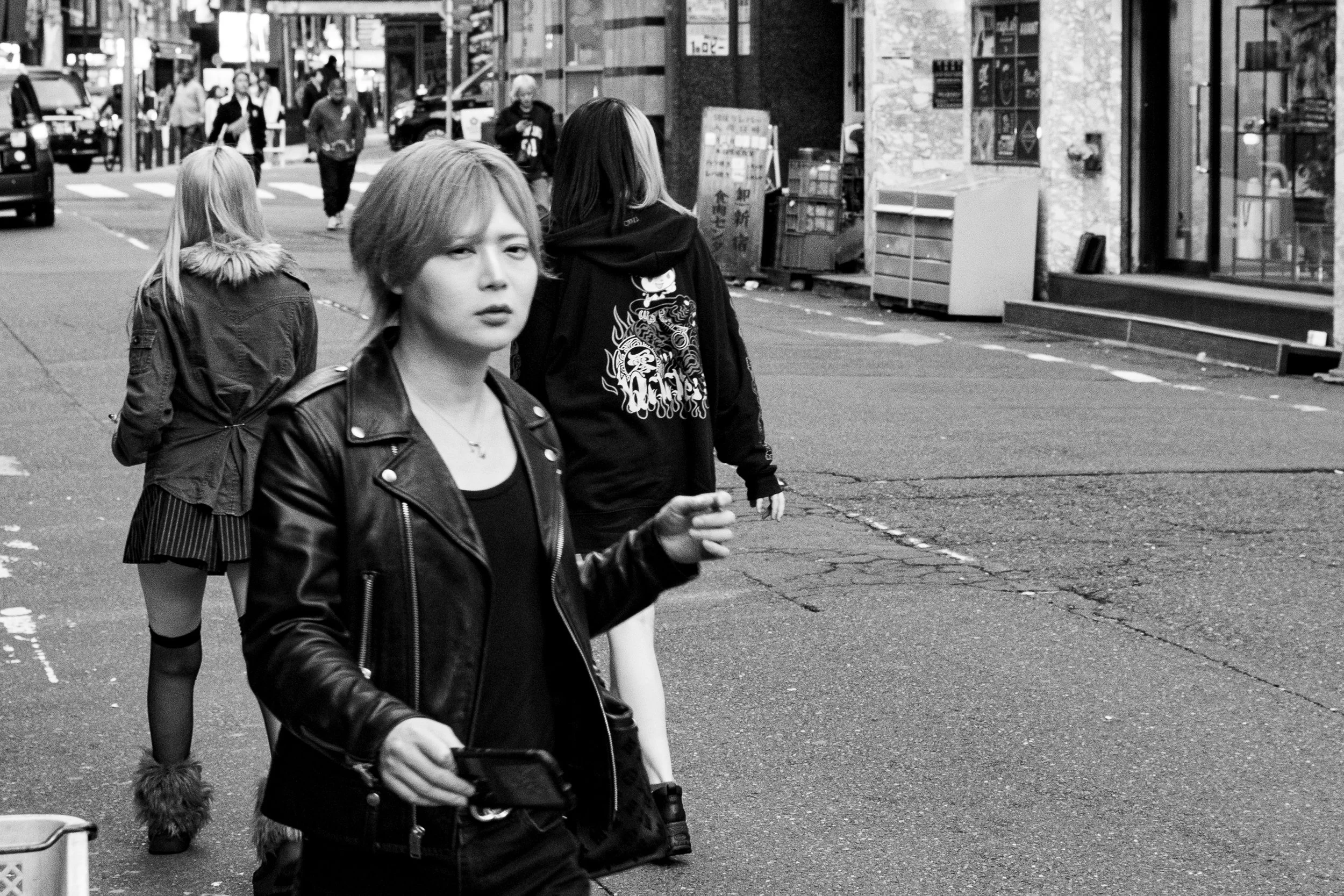 Black and white photo of three women walking on a city sidewalk, with buildings and pedestrians in the background.