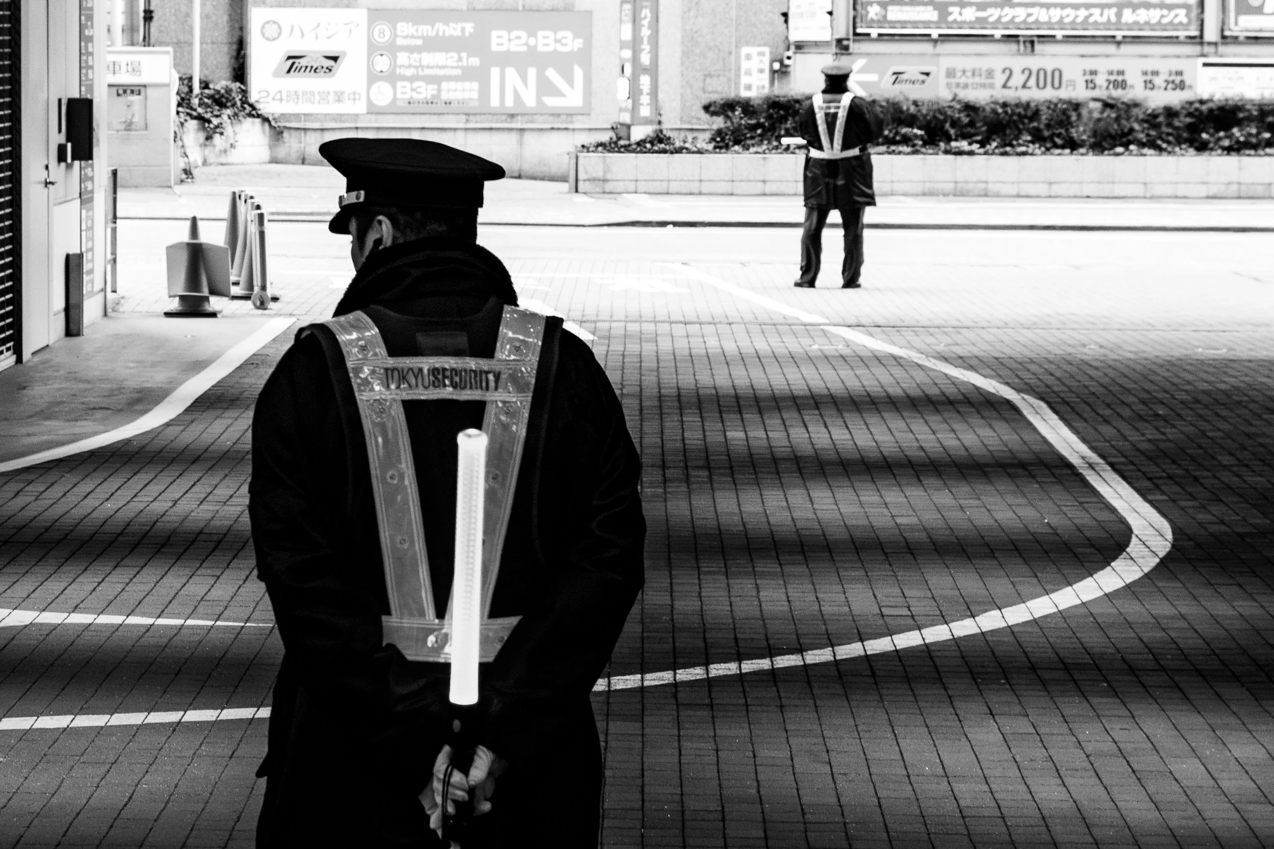 A police officer standing with his back to the camera, in a high-visibility vest, holding a traffic control bat. In the background, another officer is positioned further down the street, also with a bat, in an urban setting.