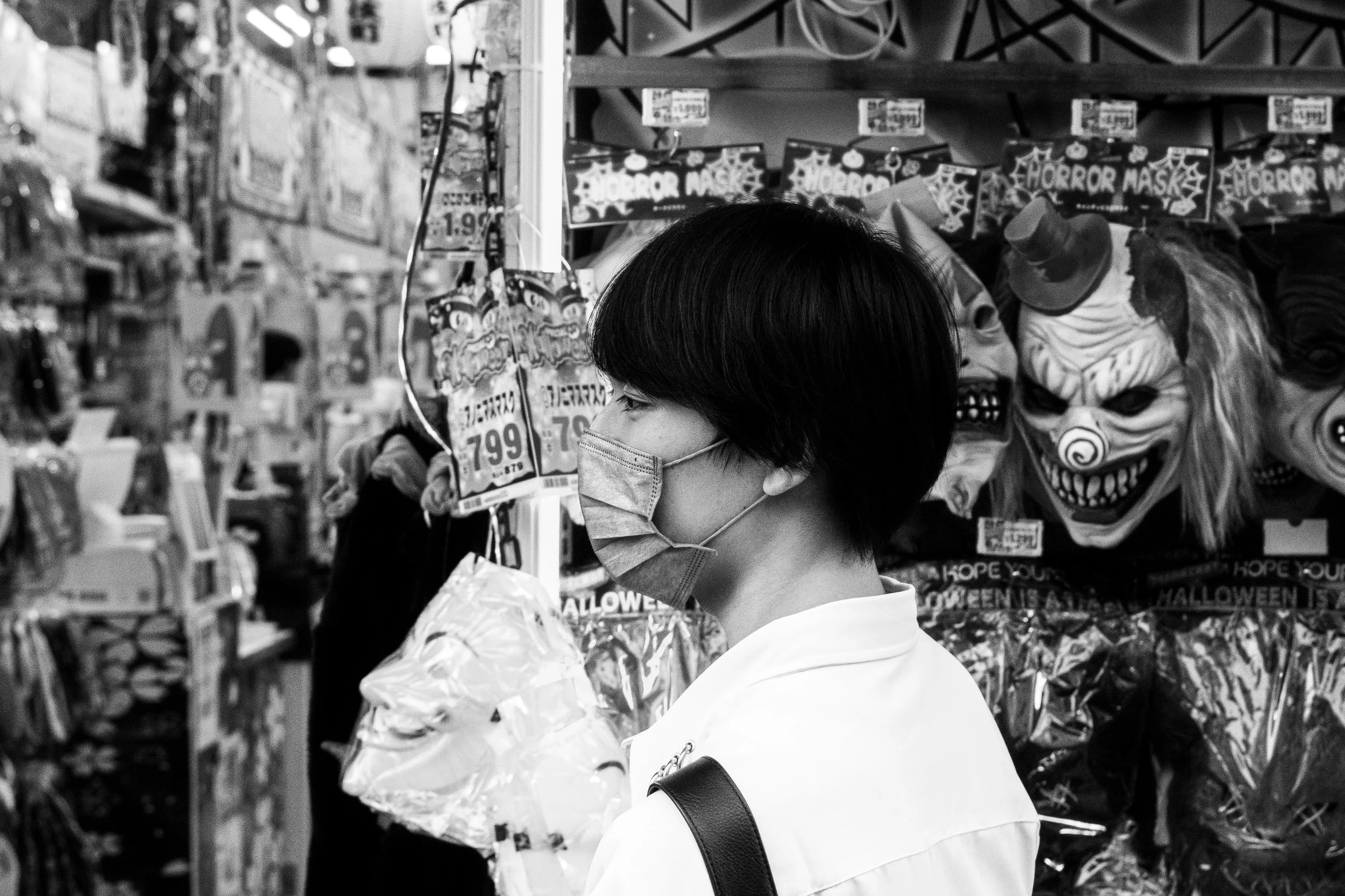 A person wearing a face mask shopping for Halloween masks at a store with Halloween costumes and masks on the shelves.