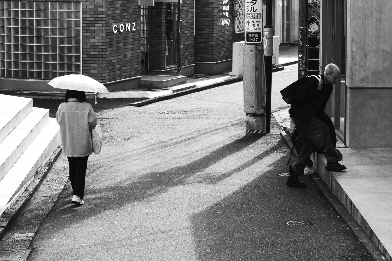 Black and white street scene with two people: a woman walking away under an umbrella and a man crouching on a sidewalk with a backpack, near a building with stairs and signs in Japanese.