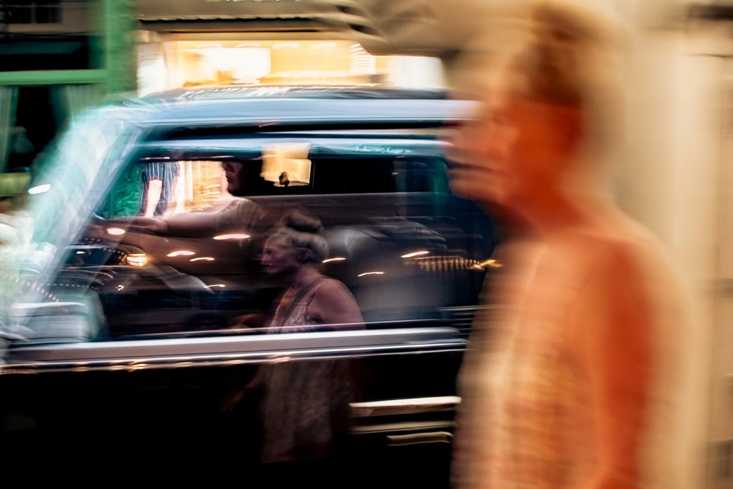 Blurry photo of a black vehicle with two people inside, a man driving and a woman sitting in the passenger seat, with a person in orange clothing walking past in the foreground.