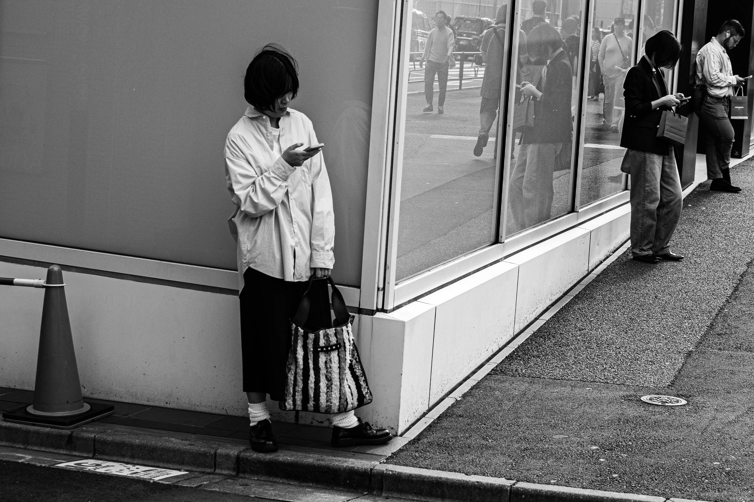 People standing outside a building, all looking at their phones, with some reflected in the glass and others walking past on the street.