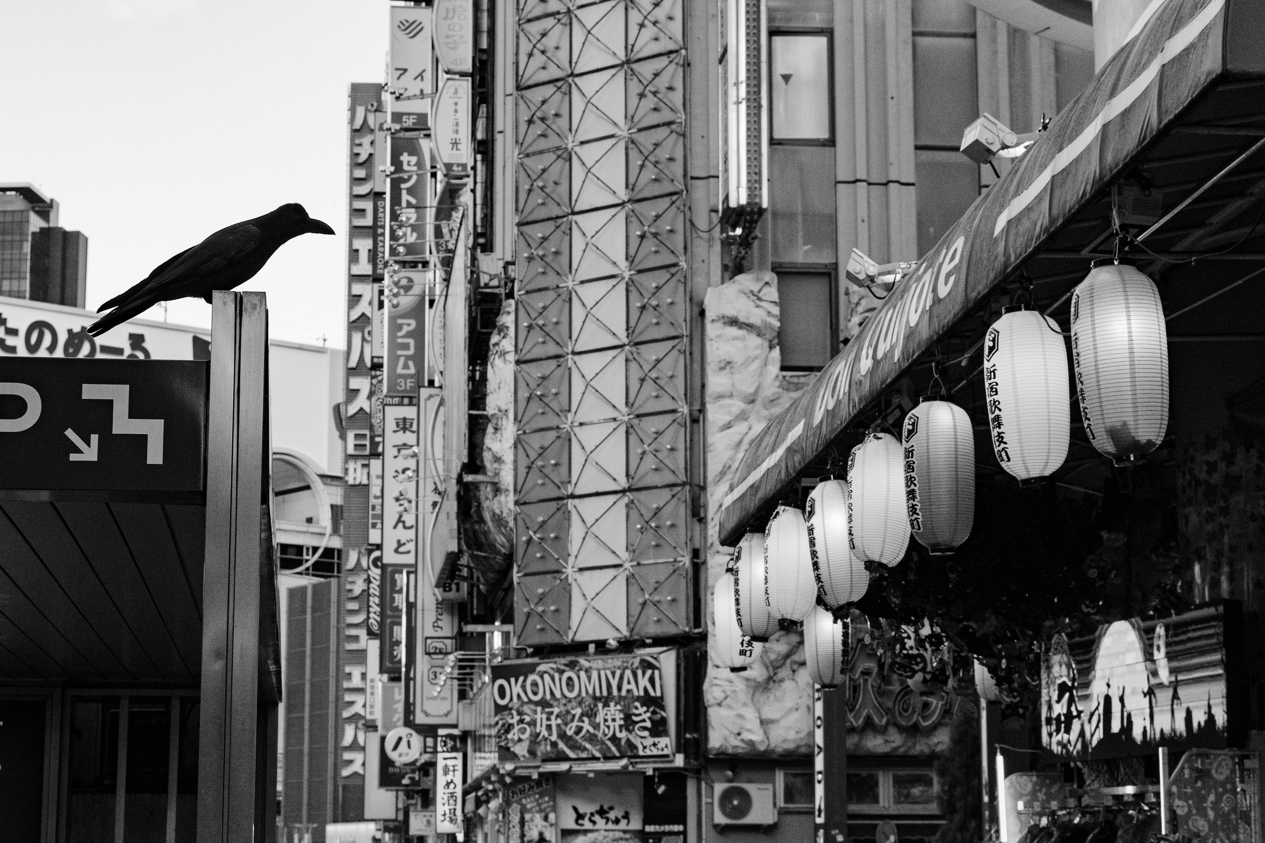 Black and white photo of a city street with hanging lanterns and various signs, including the word "OKONOMIYAKI".