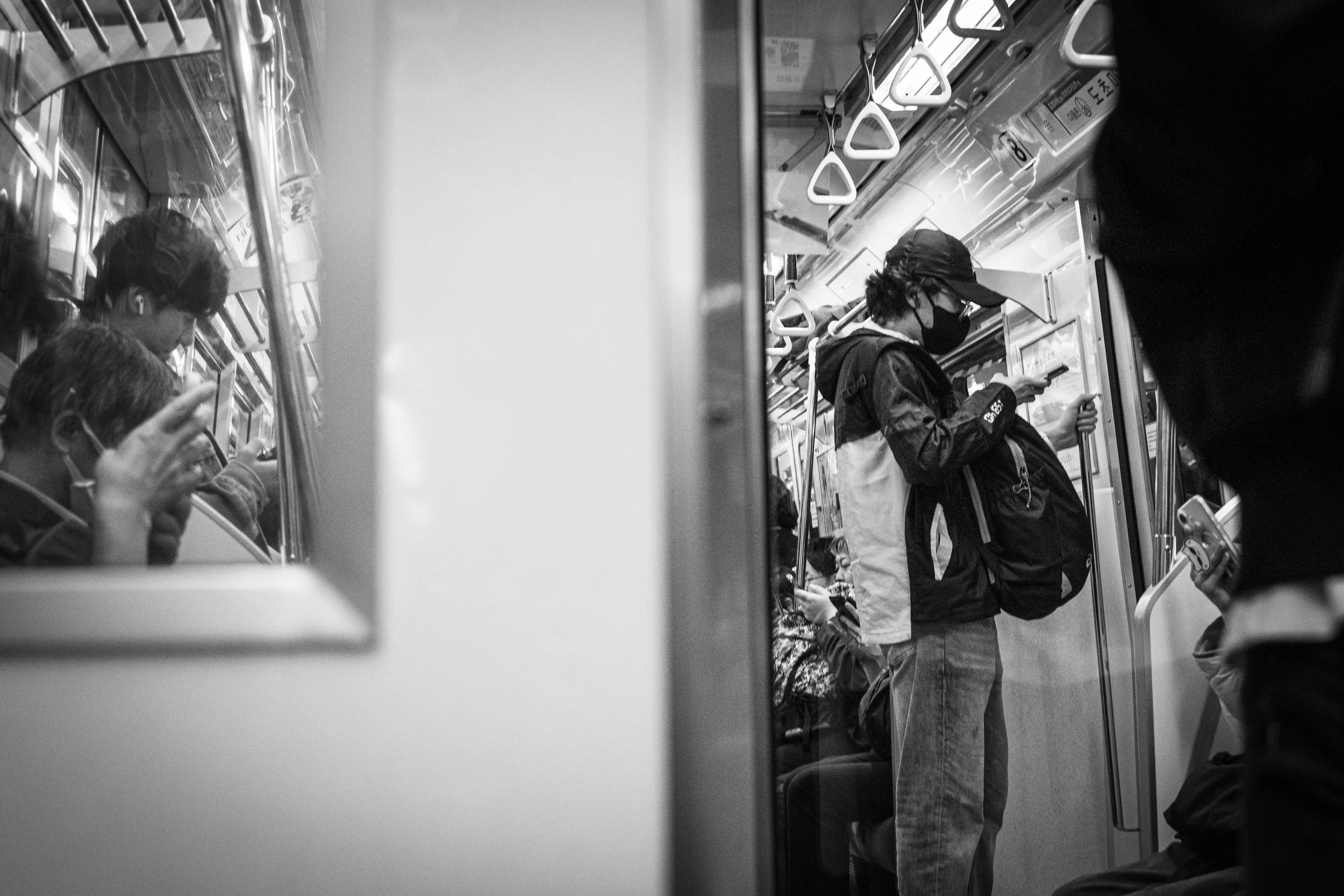 Black and white photo of a crowded subway car with passengers focused on their phones, some wearing masks, standing and sitting.
