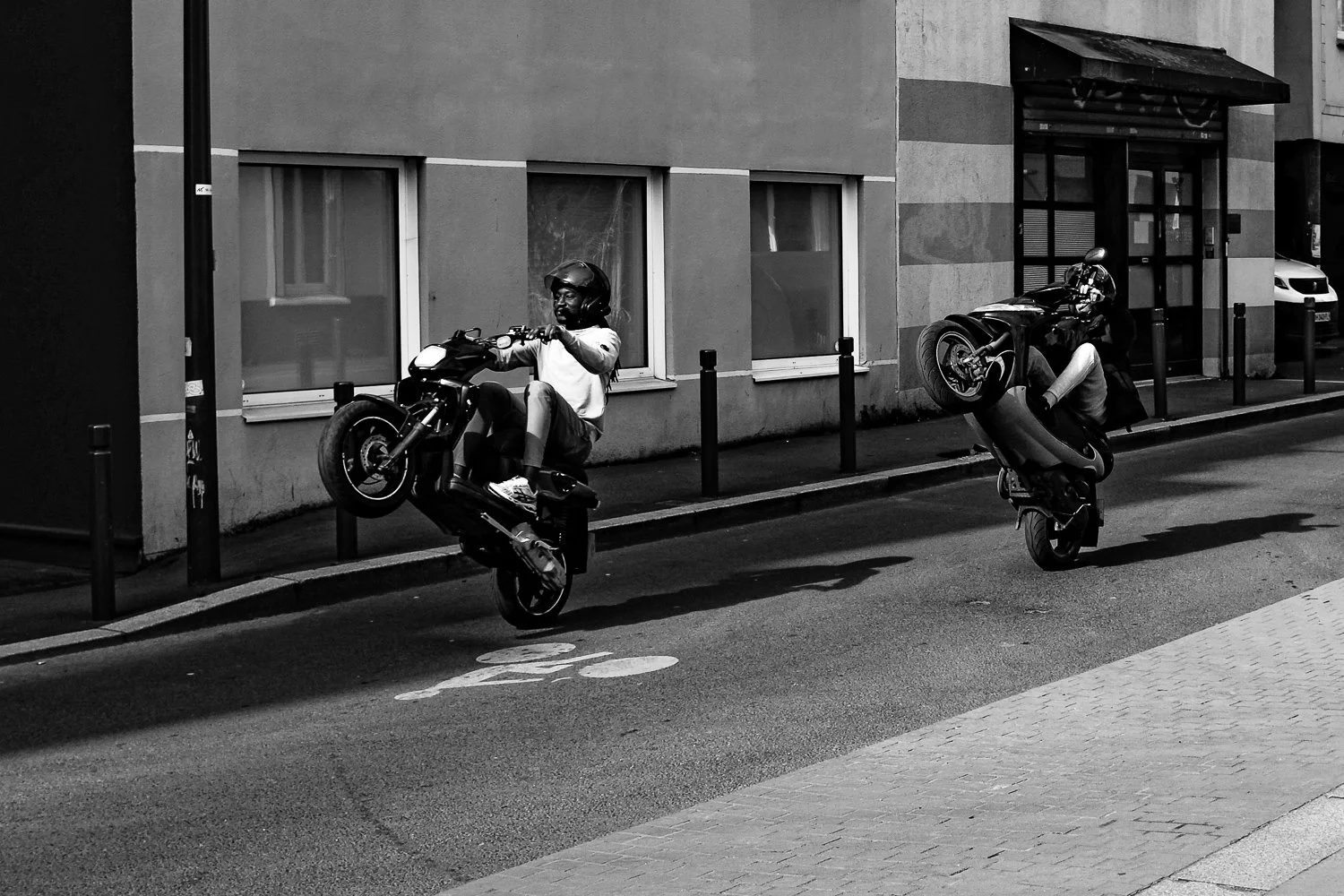 Two motorcyclists performing wheelies on a city street at night, with building windows and parked cars in the background.
