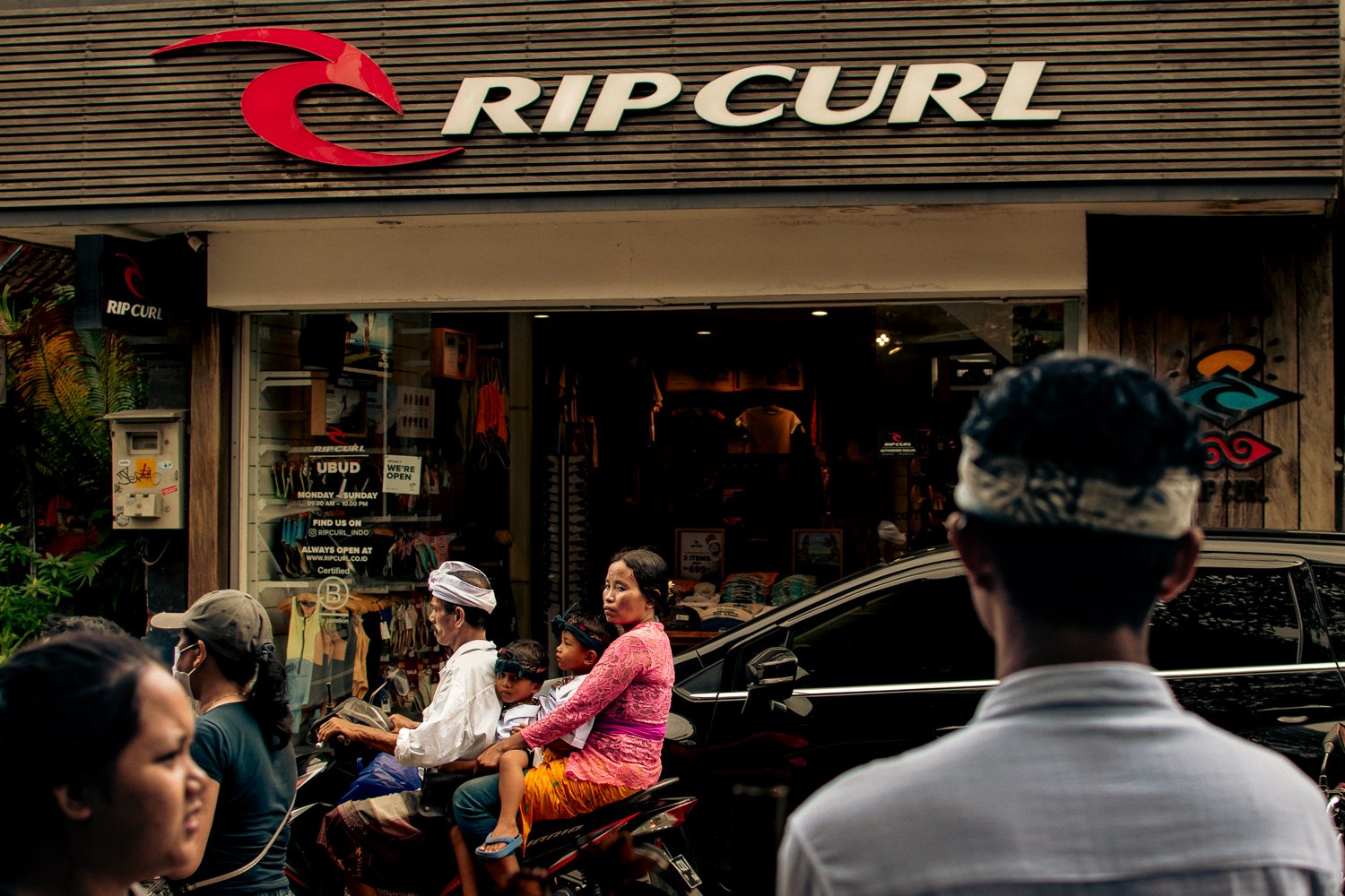 Street scene in front of a Rip Curl store with people riding motorbikes and cars passing by.