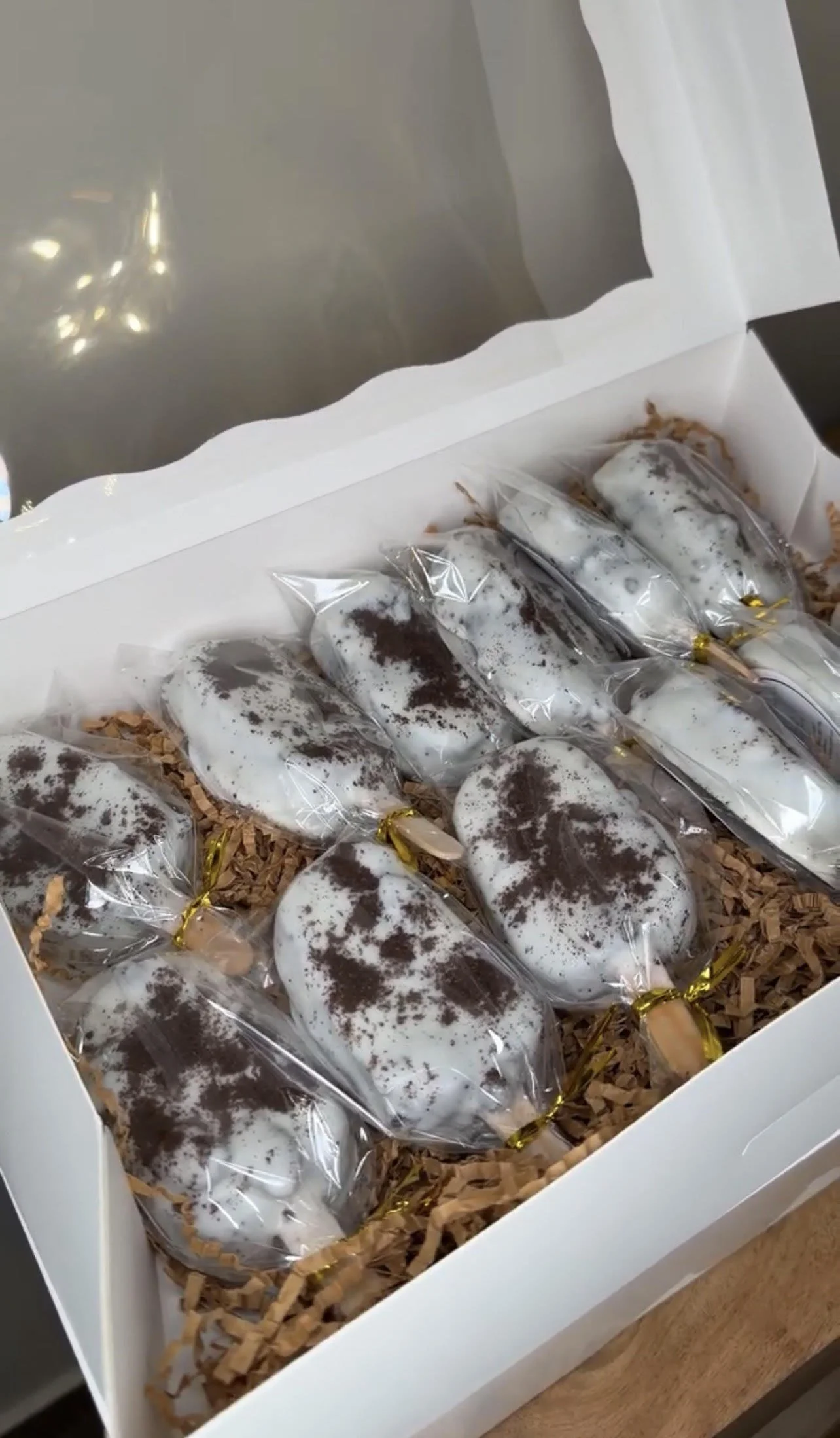 A box of cookies wrapped in clear plastic, with powdered sugar and cocoa powder on top, arranged on shredded paper inside a white box.