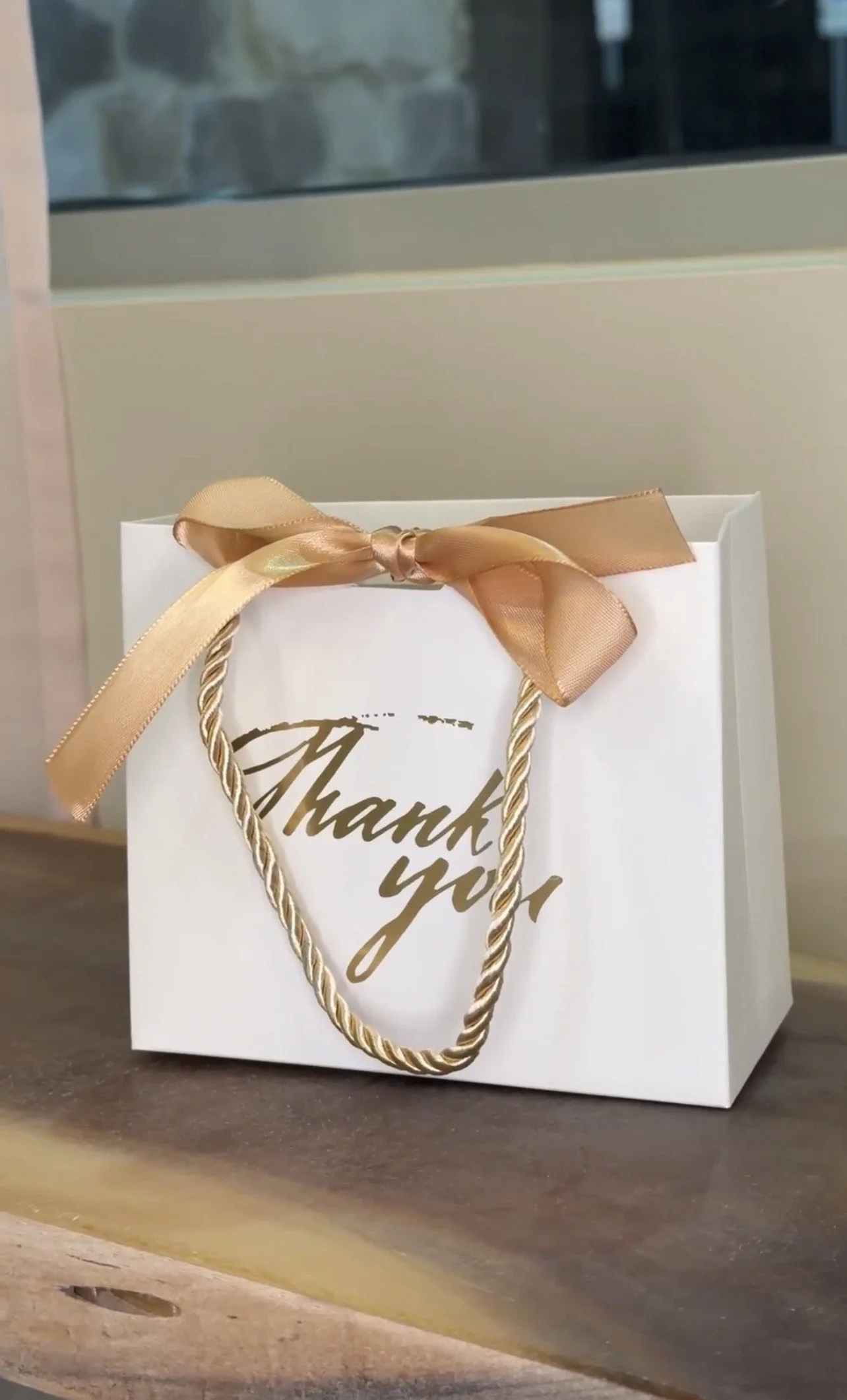 A white gift bag with gold lettering that says 'Thank you'. The bag has a gold ribbon bow on the handle and is placed on a wooden surface near a window with a stone wall visible outside.