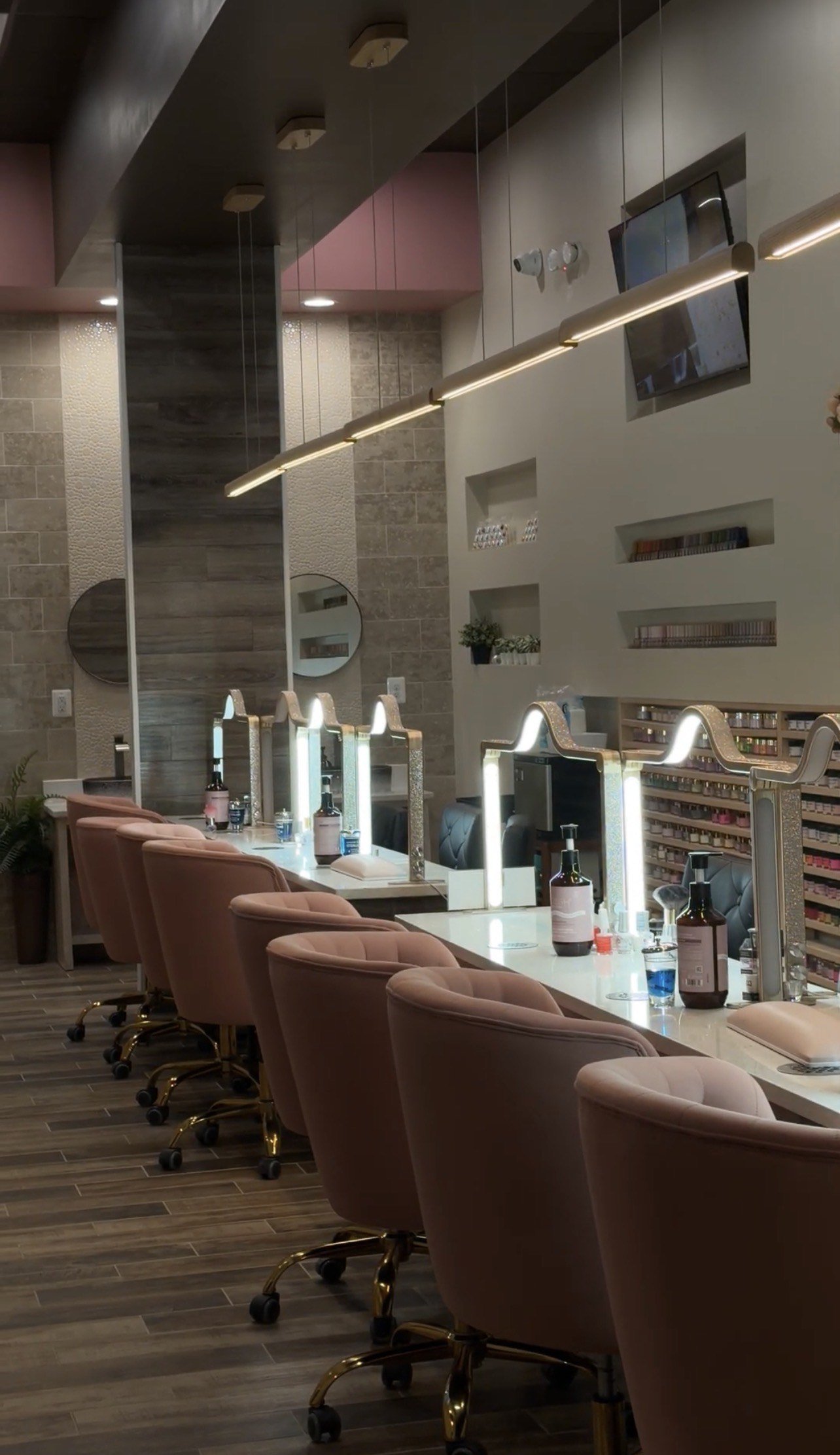Beauty salon with vanity stations, pink chairs, illuminated mirrors, nail polish shelves, and a wall-mounted TV.