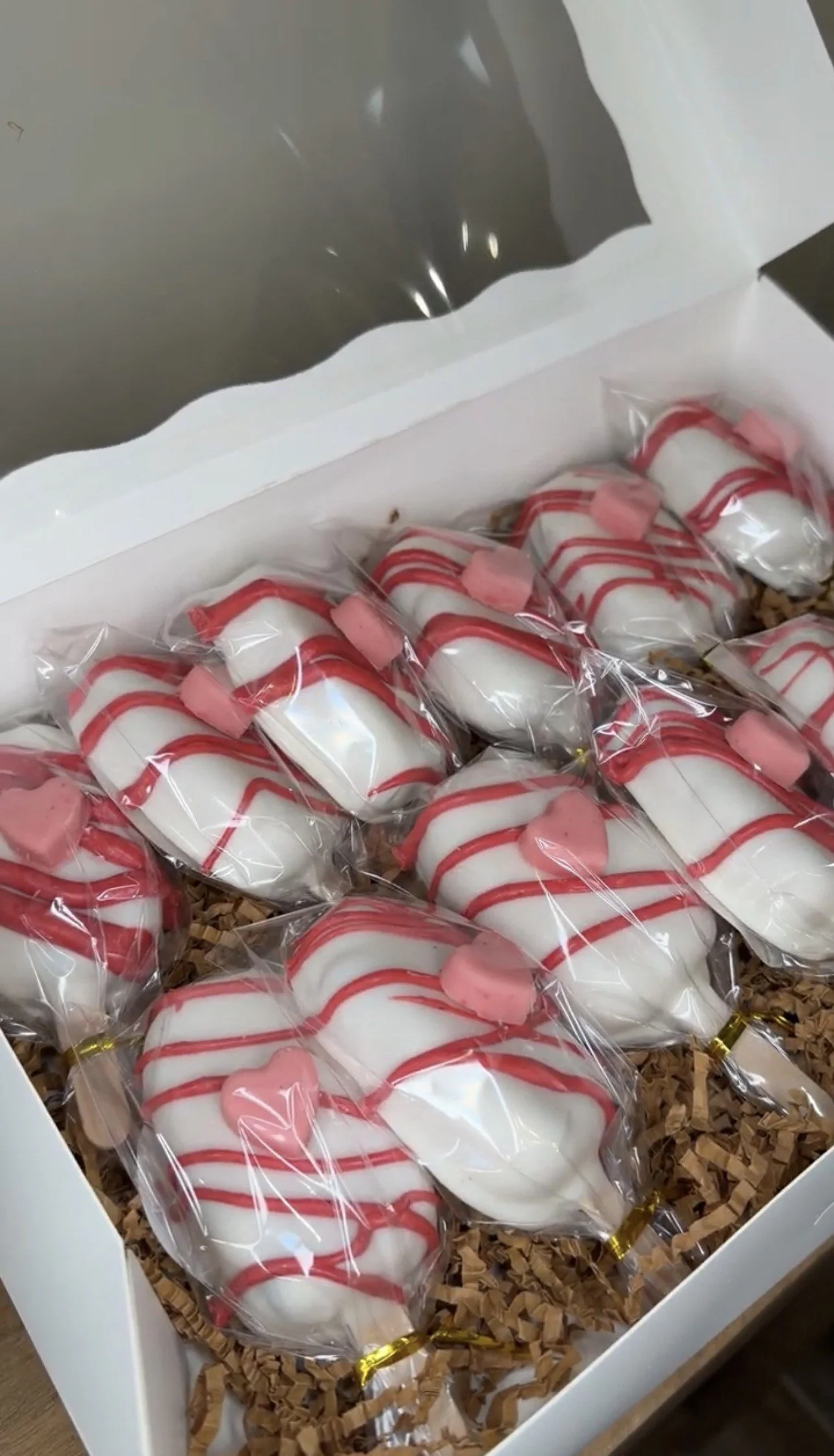 A box filled with individually wrapped, white, heart-shaped sugar cookies decorated with red icing stripes and pink heart decorations, placed on crumpled brown paper shred inside a white box.