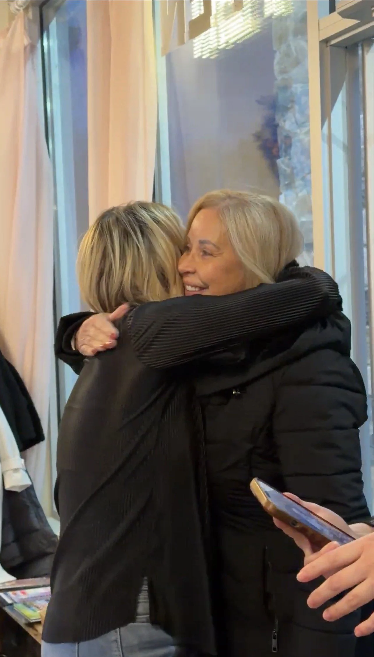 Two women are hugging warmly indoors, one facing away from the camera and the other smiling with her eyes closed.