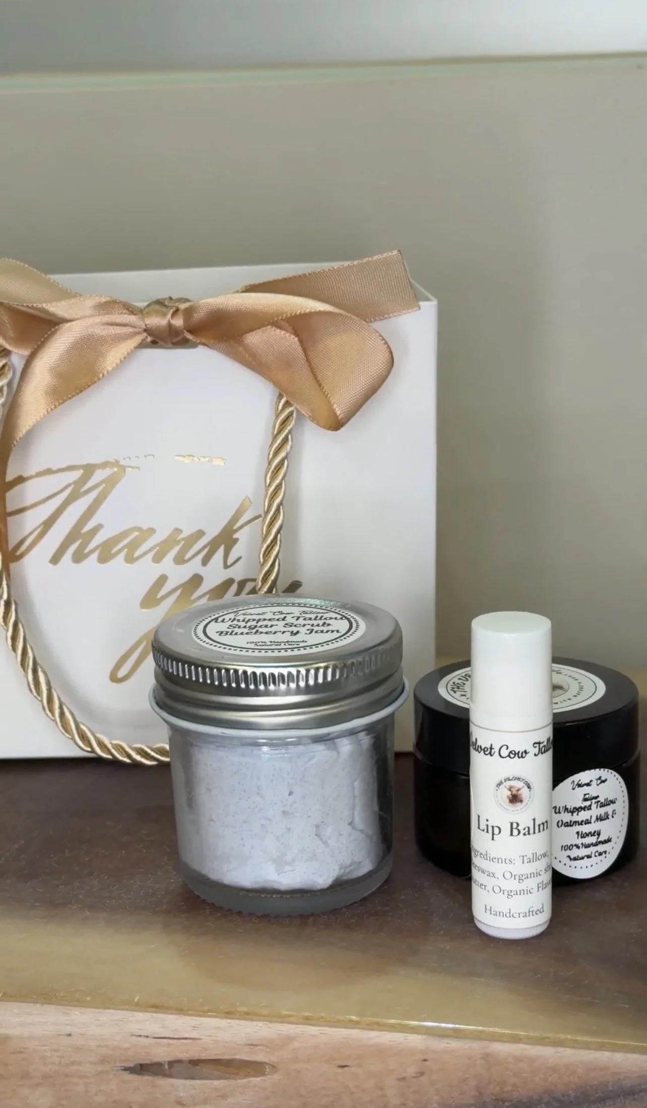 A gift bag with a golden ribbon and bow labeled 'Thank you' next to jars of body scrub and lip balm. The body scrub jar is labeled 'Whipped Fallow Sugar Scrub Blueberry Jam' and the lip balm tube is labeled 'Lip Balm' with Tallow, Shea Butter ingredients.