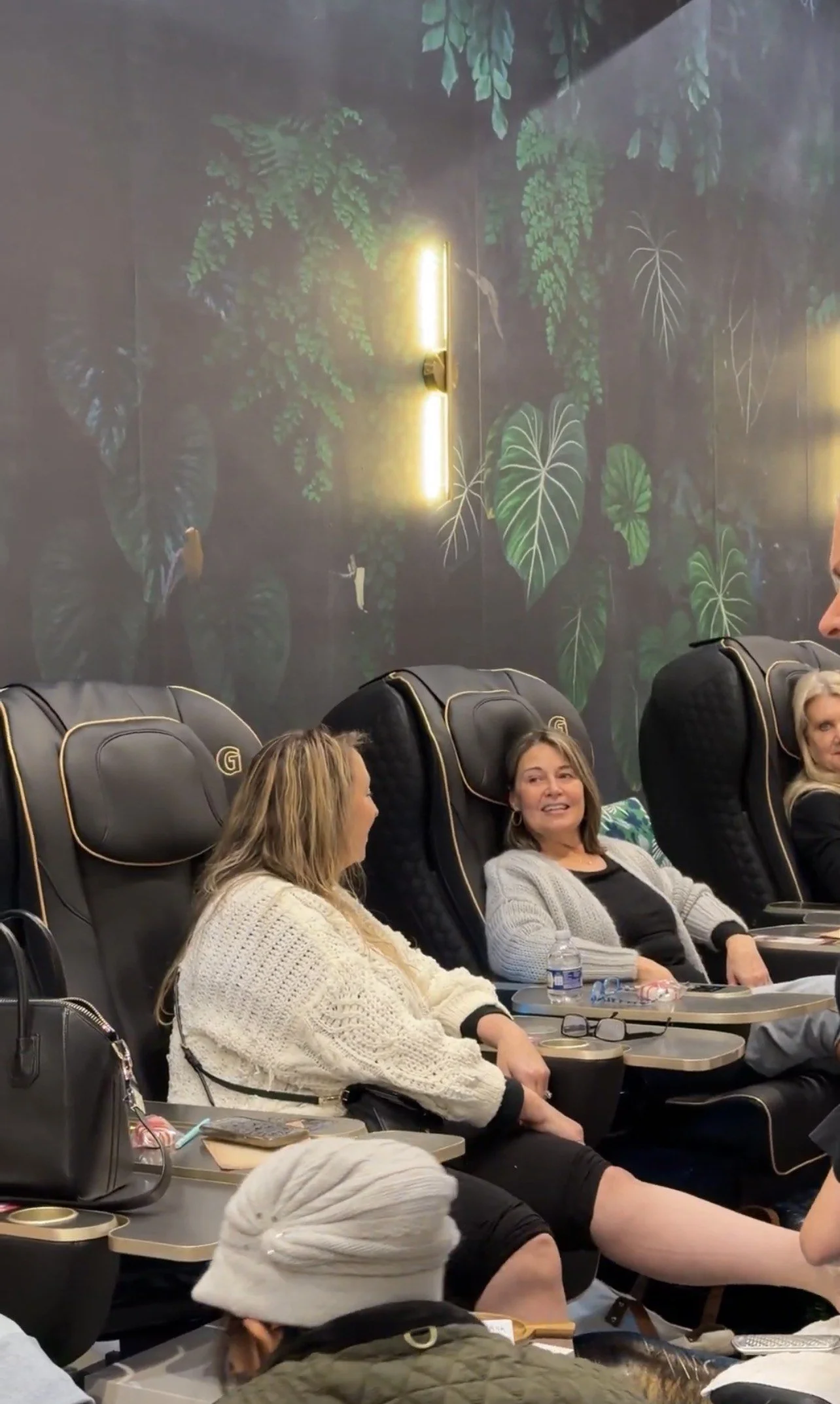 Three women sitting in massage chairs in a room with a dark green leaf-patterned wall and gold-accented lighting.