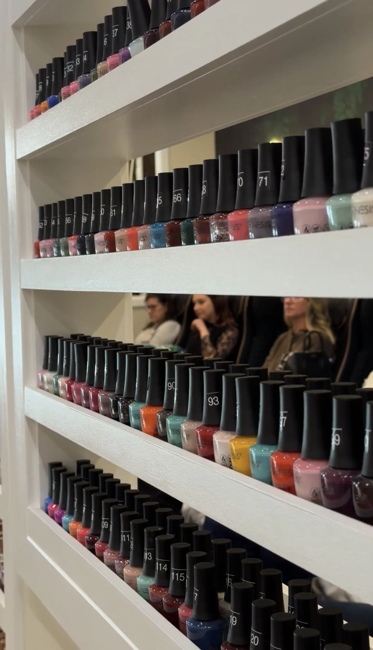 A white nail polish display shelf with multiple rows of colorful nail polish bottles at a store or salon. The bottles are organized by color and numbered on the caps.