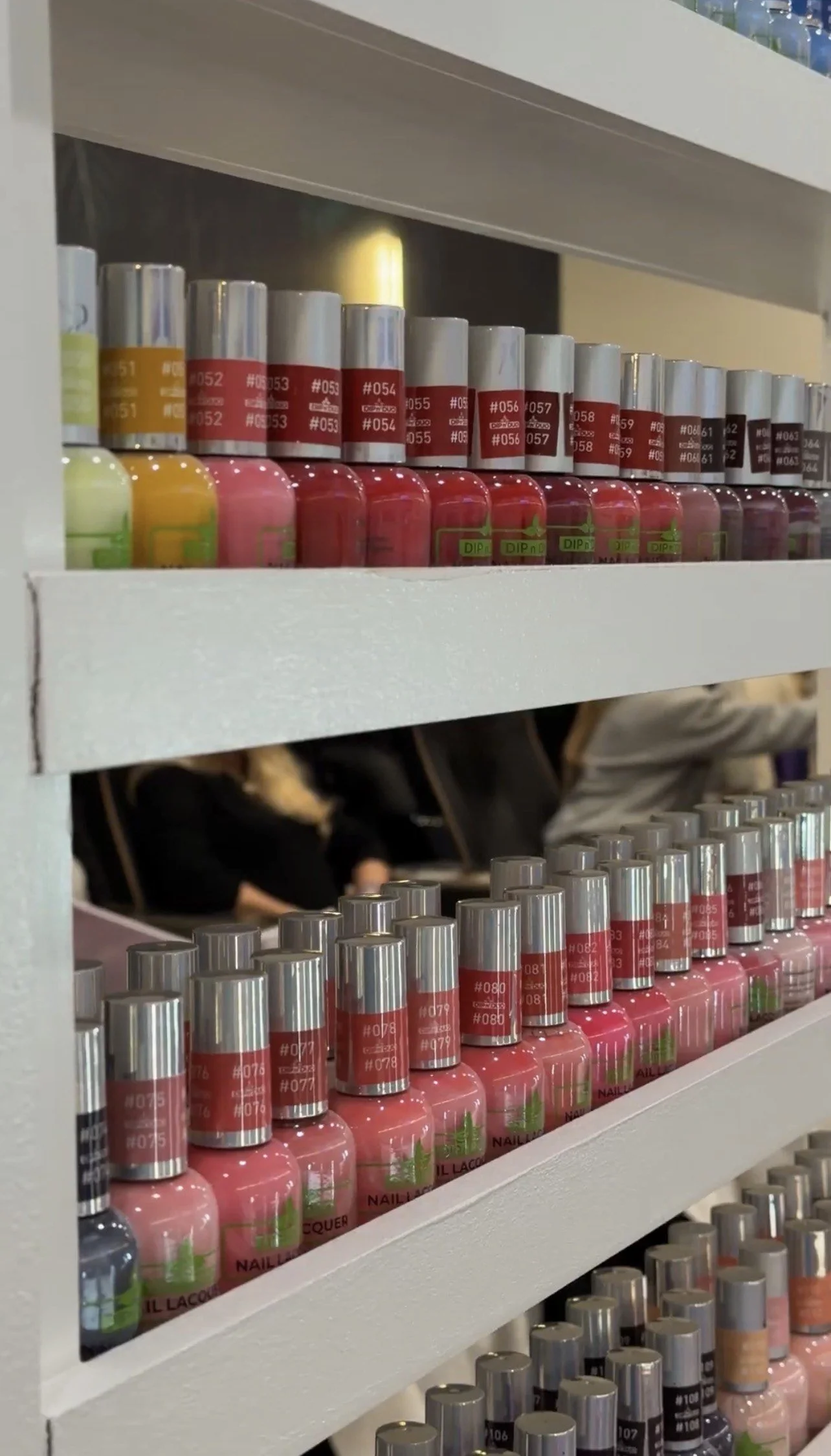 Close-up of shelves filled with colorful bottles of nail polish, arranged by color, in a salon or store. The top shelf has yellow, pink, red, and dark shades; the middle shelf has pinks and reds; and the bottom shelf has various shades of pink and beige.