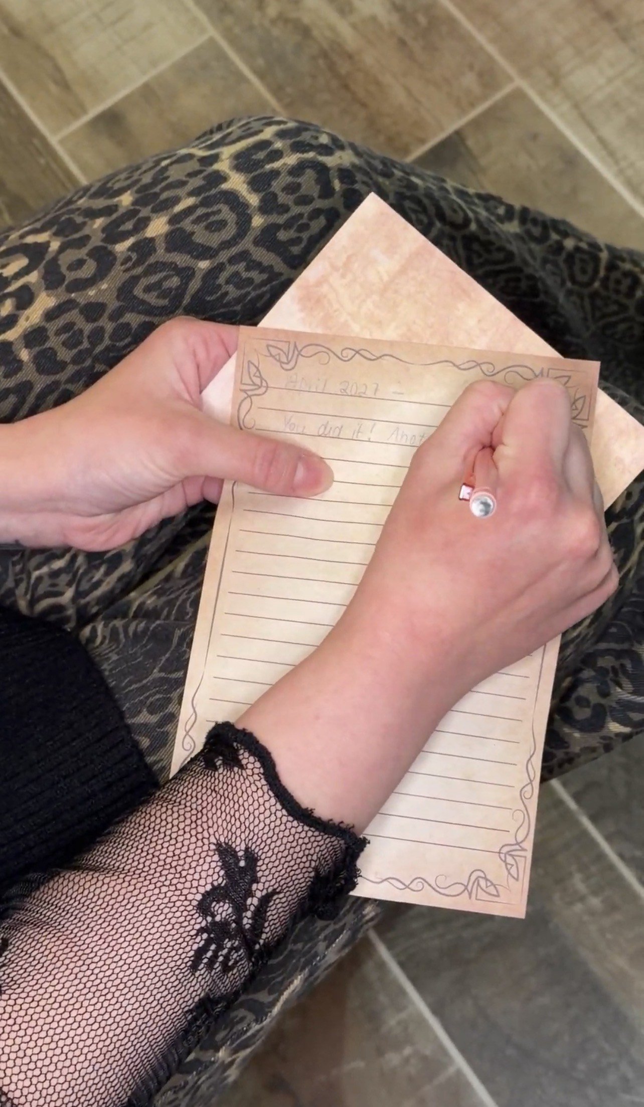 A person sitting on a patterned seat, writing a note on a decorative paper notepad with a pen. The person is wearing a black long sleeve with lace at the wrist. The background shows tiled flooring.