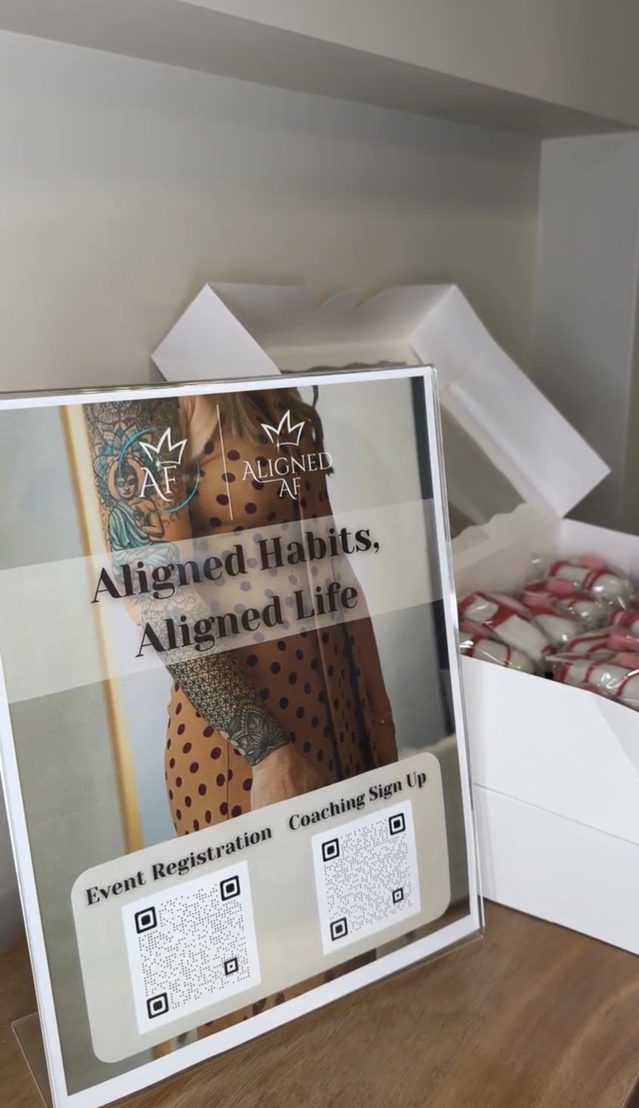 Sign promoting a coaching event with a woman's tattooed arm in the background, QR codes for event registration and coaching sign-up, and a box of small wrapped items behind the sign.