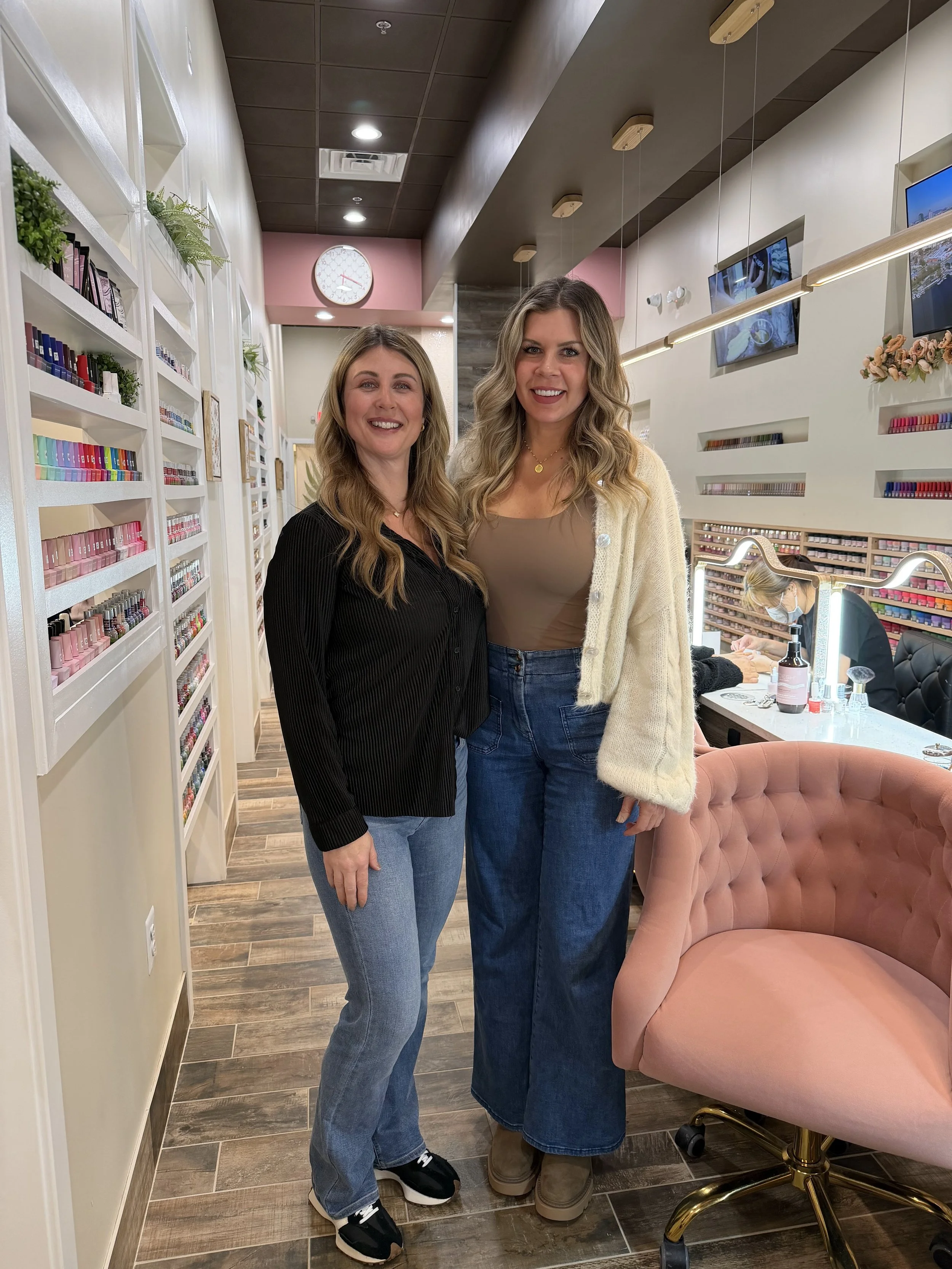 Two women smiling and standing together in a nail salon with shelves of nail polish bottles, a pink velvet chair, and a mirror with a nail technician at work in the background.