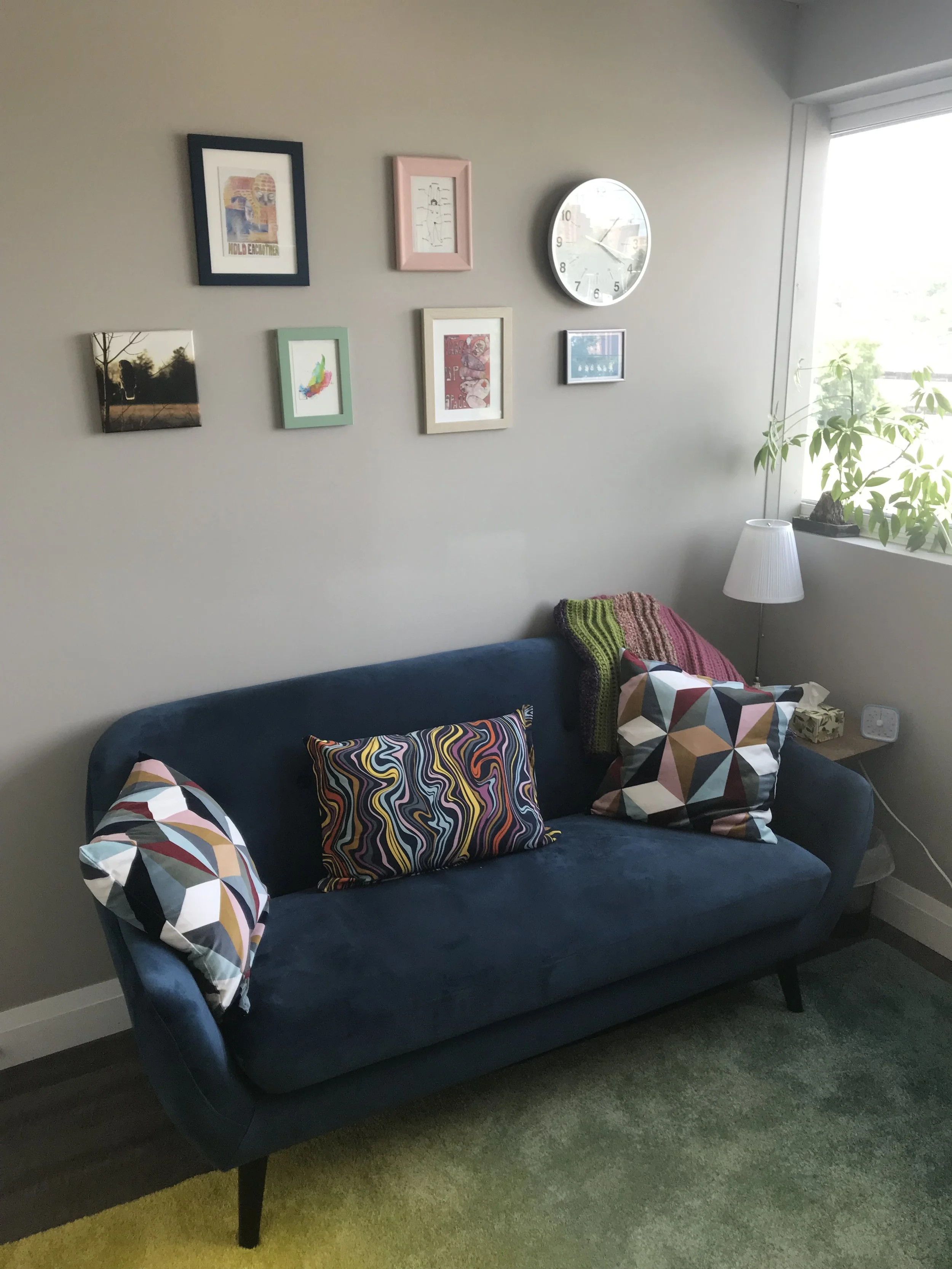 A blue velvet couch with colourful pillows rests against a beige wall with a photo gallery of art, images, and a clock above it.