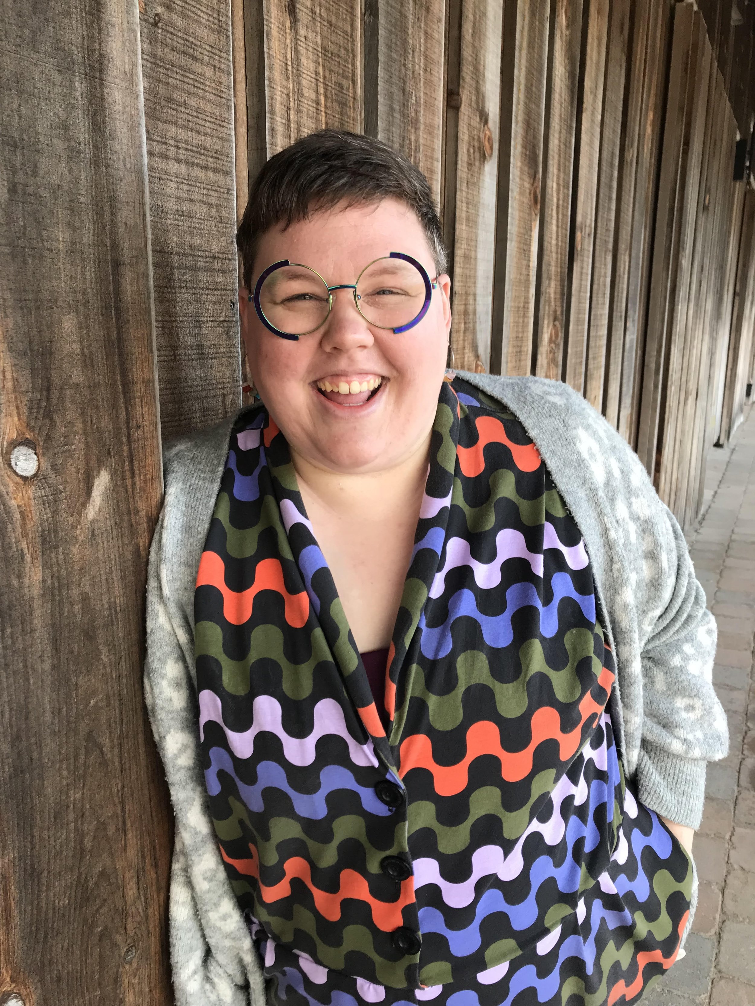 Becca is a smiling person with short dark hair, wearing round glasses, a colorful patterned outfit, and a gray cardigan, standing against a wooden wall.