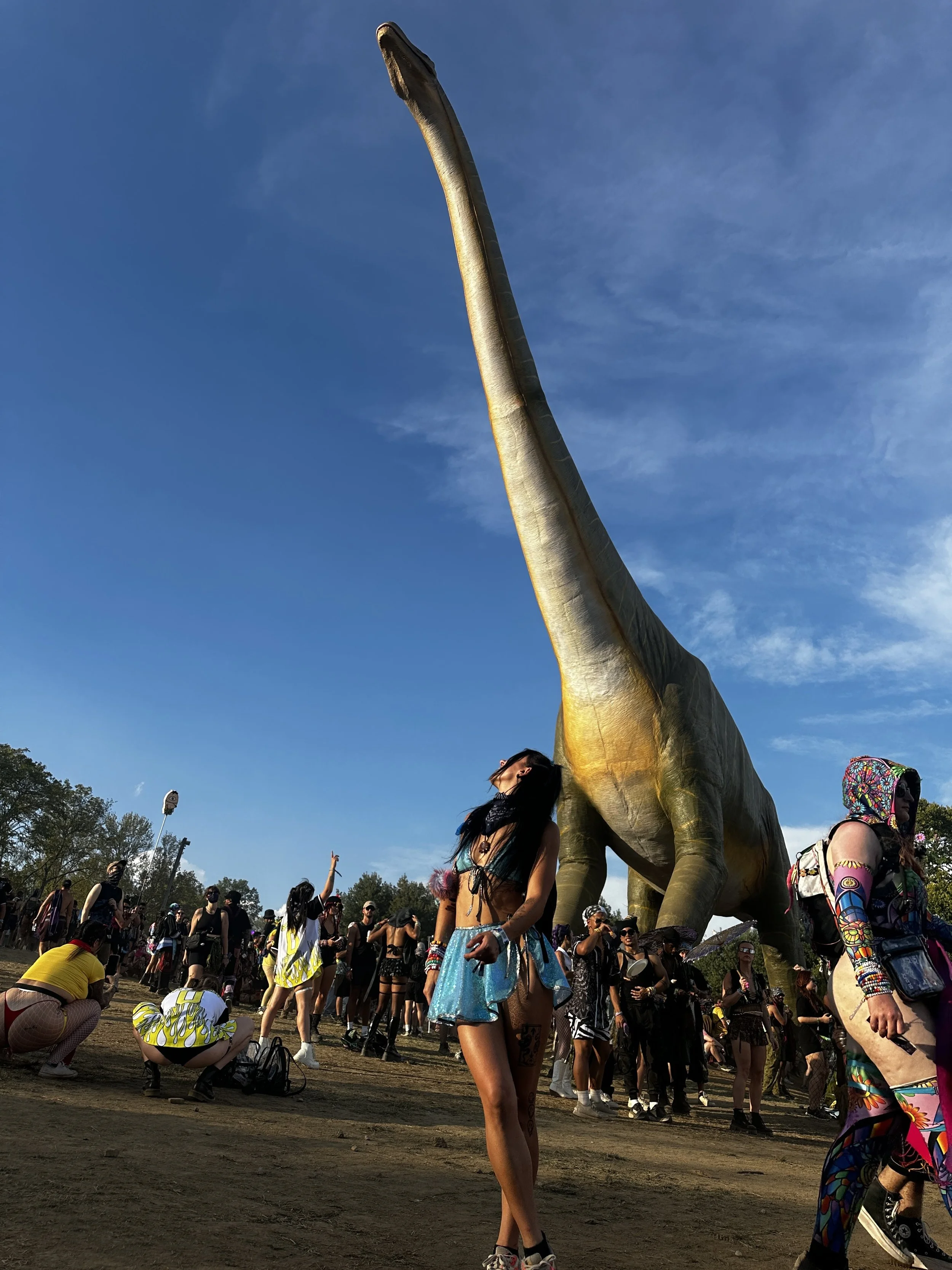 People at a festival with a large dinosaur statue in the background, some people are dancing and some are taking photos, dressed in colorful festival outfits.