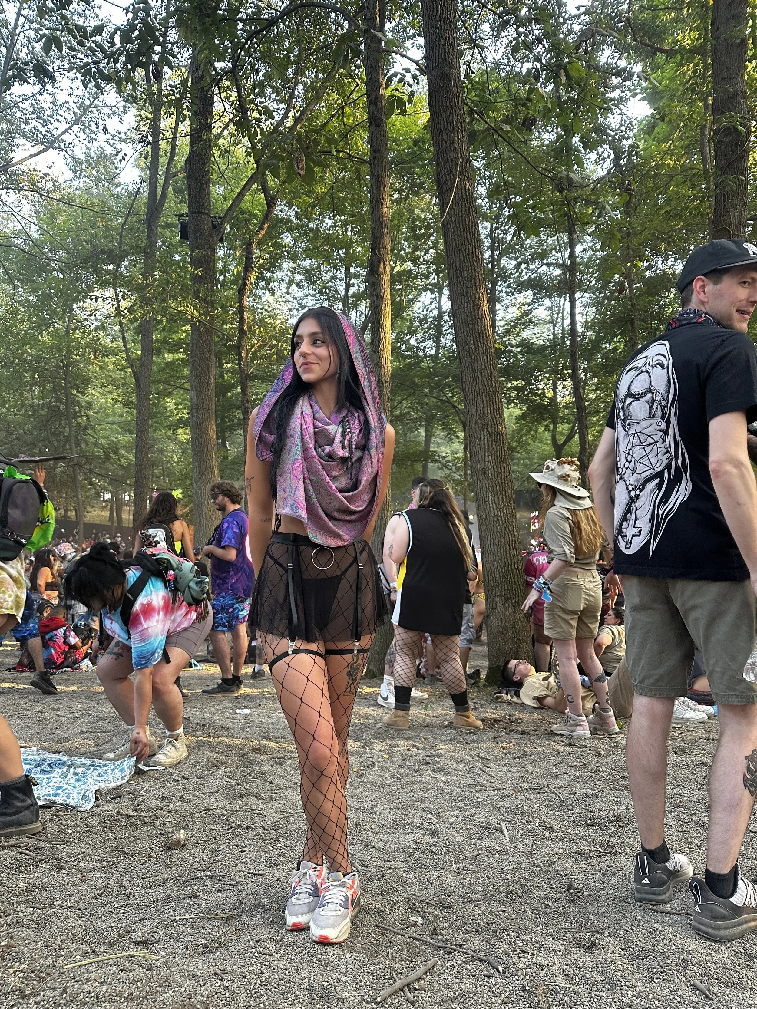 A young woman standing in a wooded outdoor area at what appears to be a festival. She is wearing a pink and purple patterned headscarf, a black sheer skirt with fishnet stockings underneath, and sneakers. Multiple people are around her, some sitting on the ground and others standing, with trees and a crowd in the background.