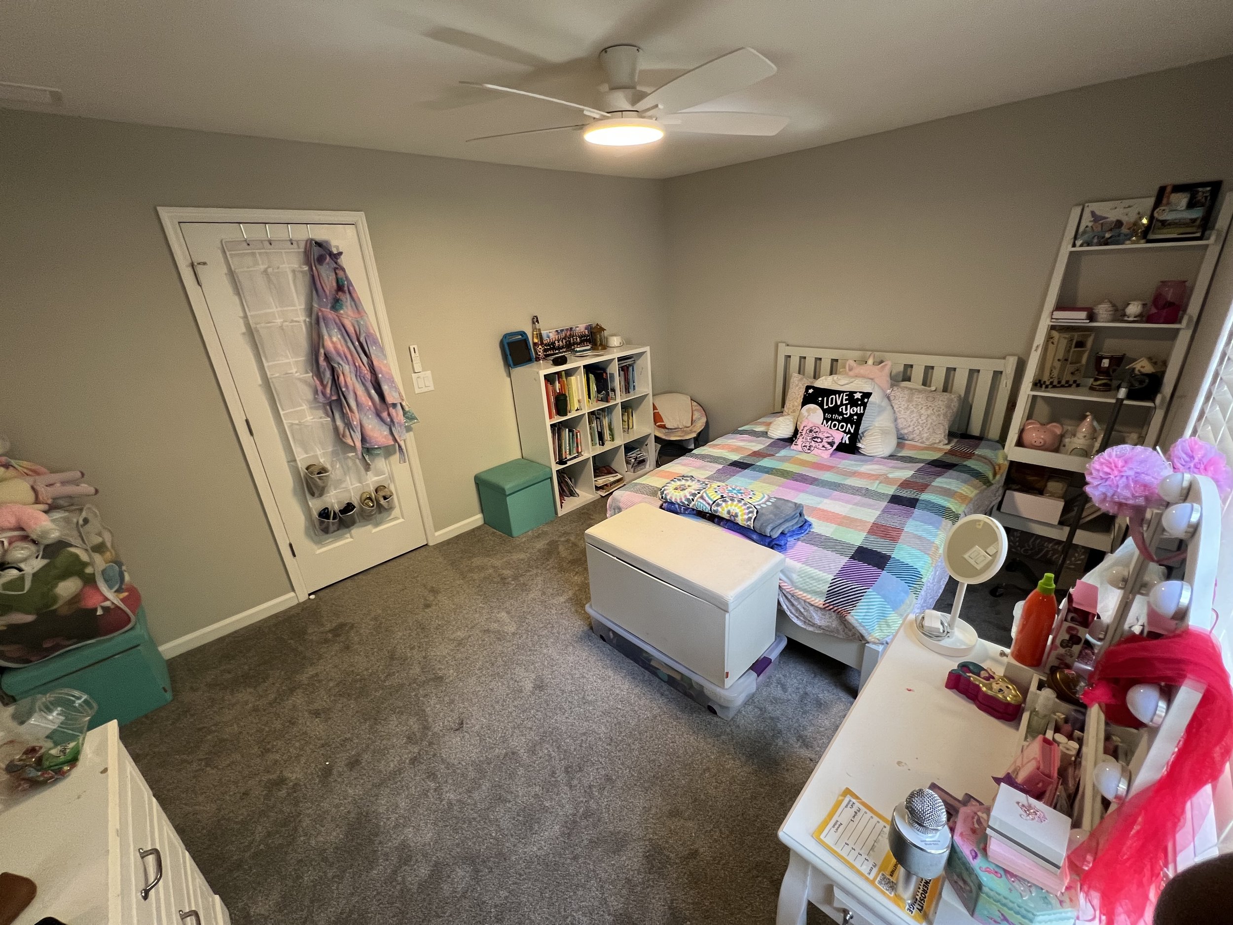 A cozy bedroom with a bed, bookshelves, a ceiling fan, and various personal items and decorations.