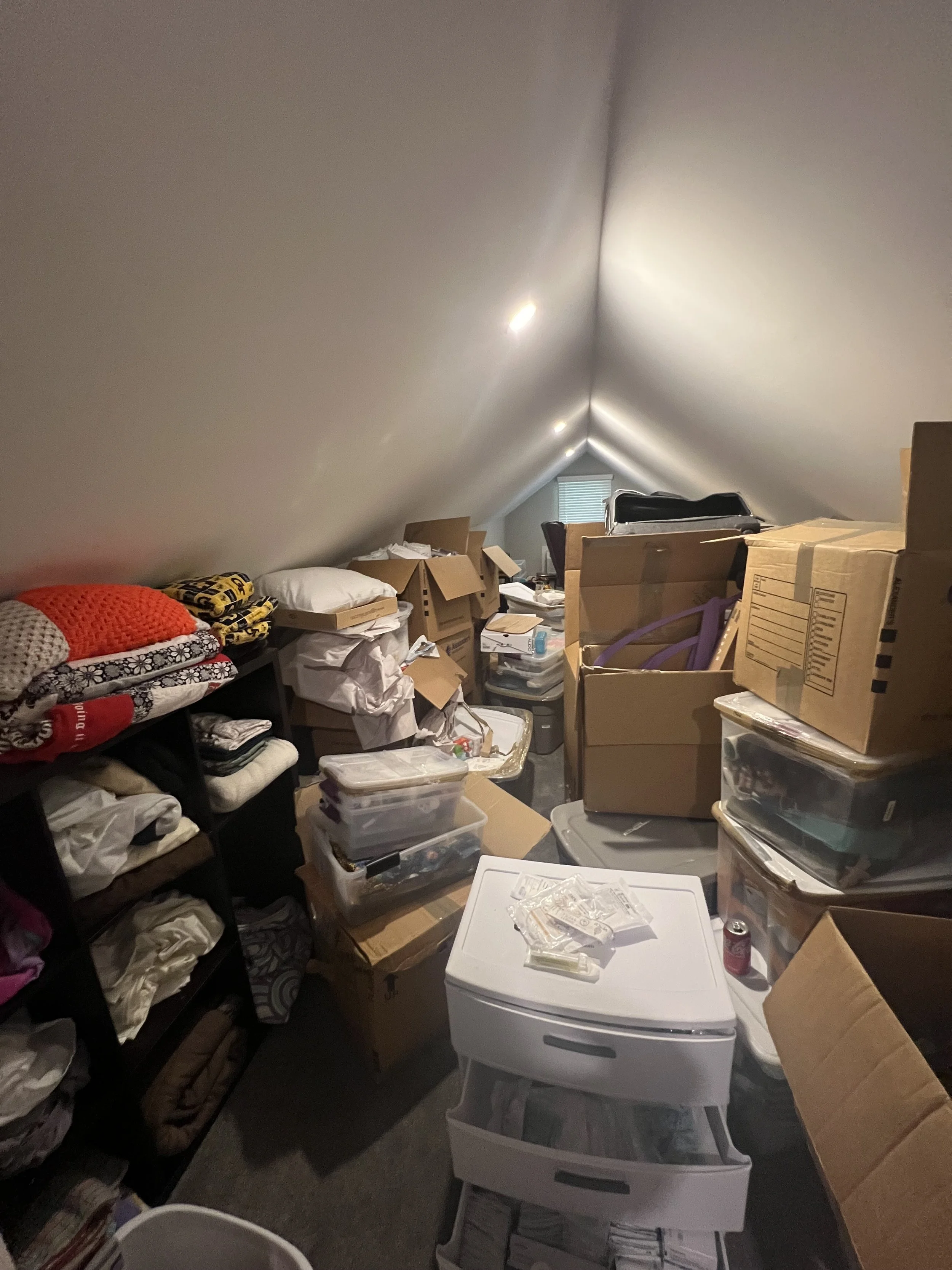Cluttered attic storage space with boxes, plastic containers, shelves, and miscellaneous items.