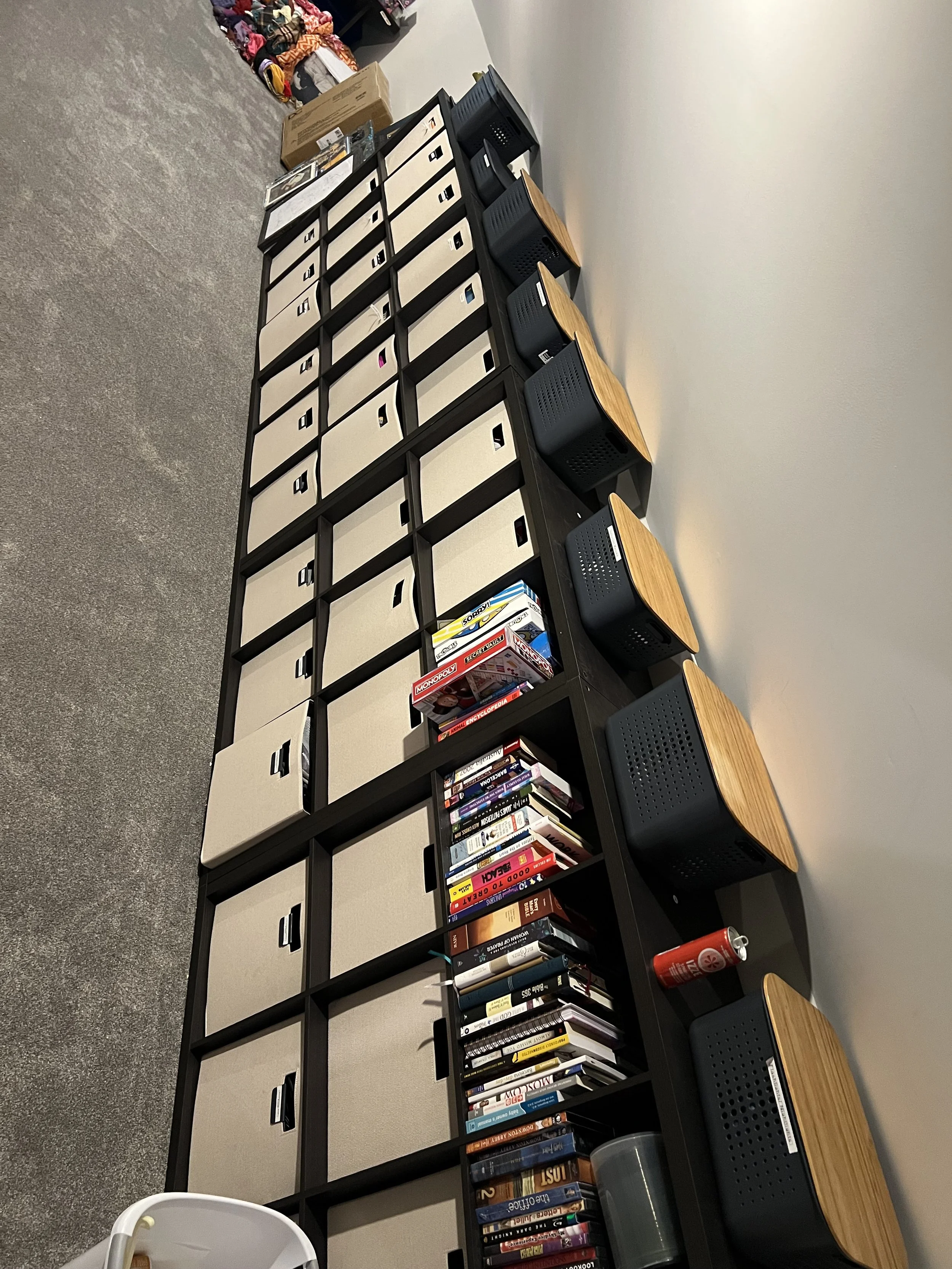Bookshelf with compartments, some holding books, others empty, against a wall. Several small stools are placed on top of the bookshelf. There are boxes and miscellaneous items on the floor nearby.