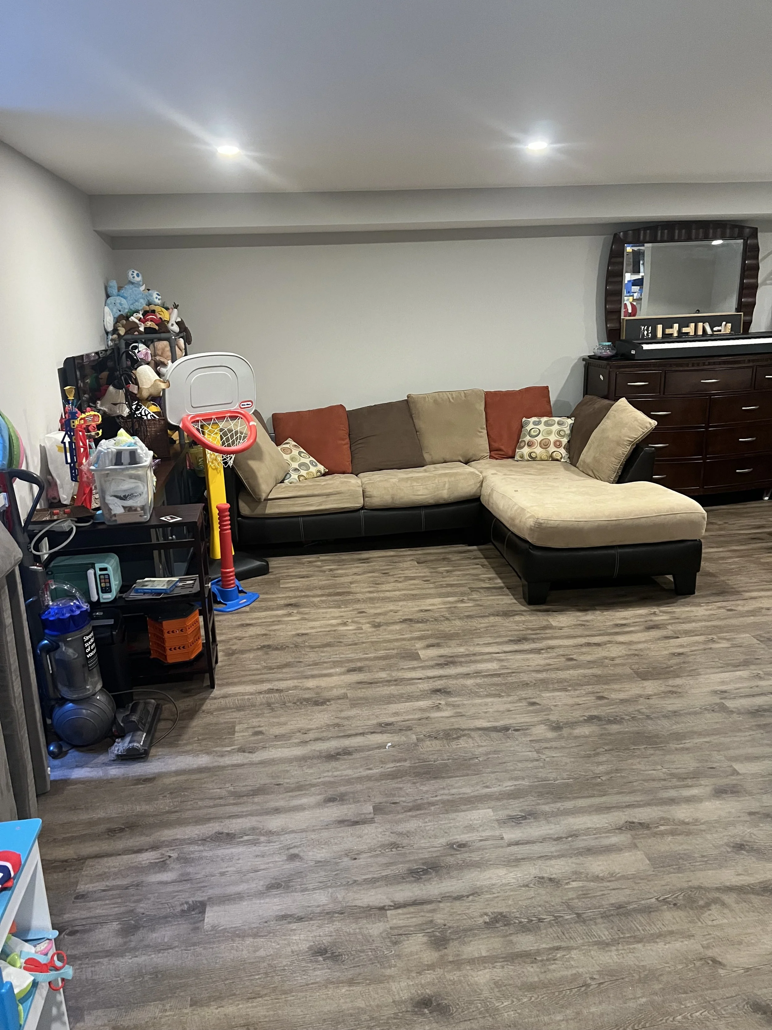 Living room with a sectional sofa, a wooden dresser with a mirror, and a shelf with toys and stuffed animals, hardwood floors, and ceiling lights.