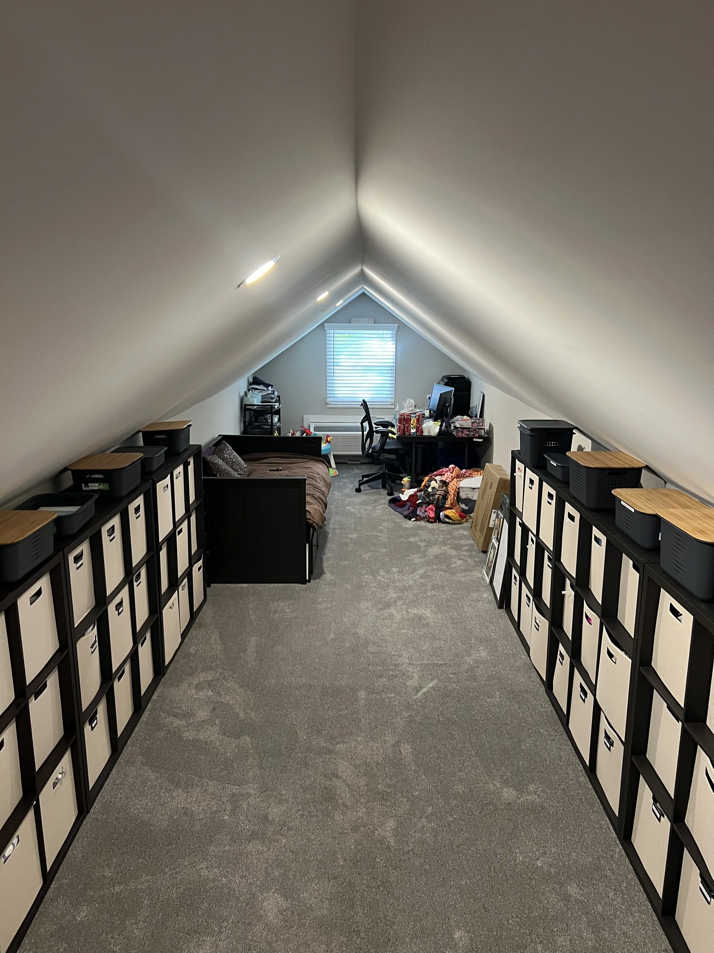 Attic room with sloped ceiling, white walls, grey carpet, black storage cubbies on both sides, small bed on the left, desk with computer and chair at the far end, clutter of clothes and boxes near the desk, window with blinds at the back.