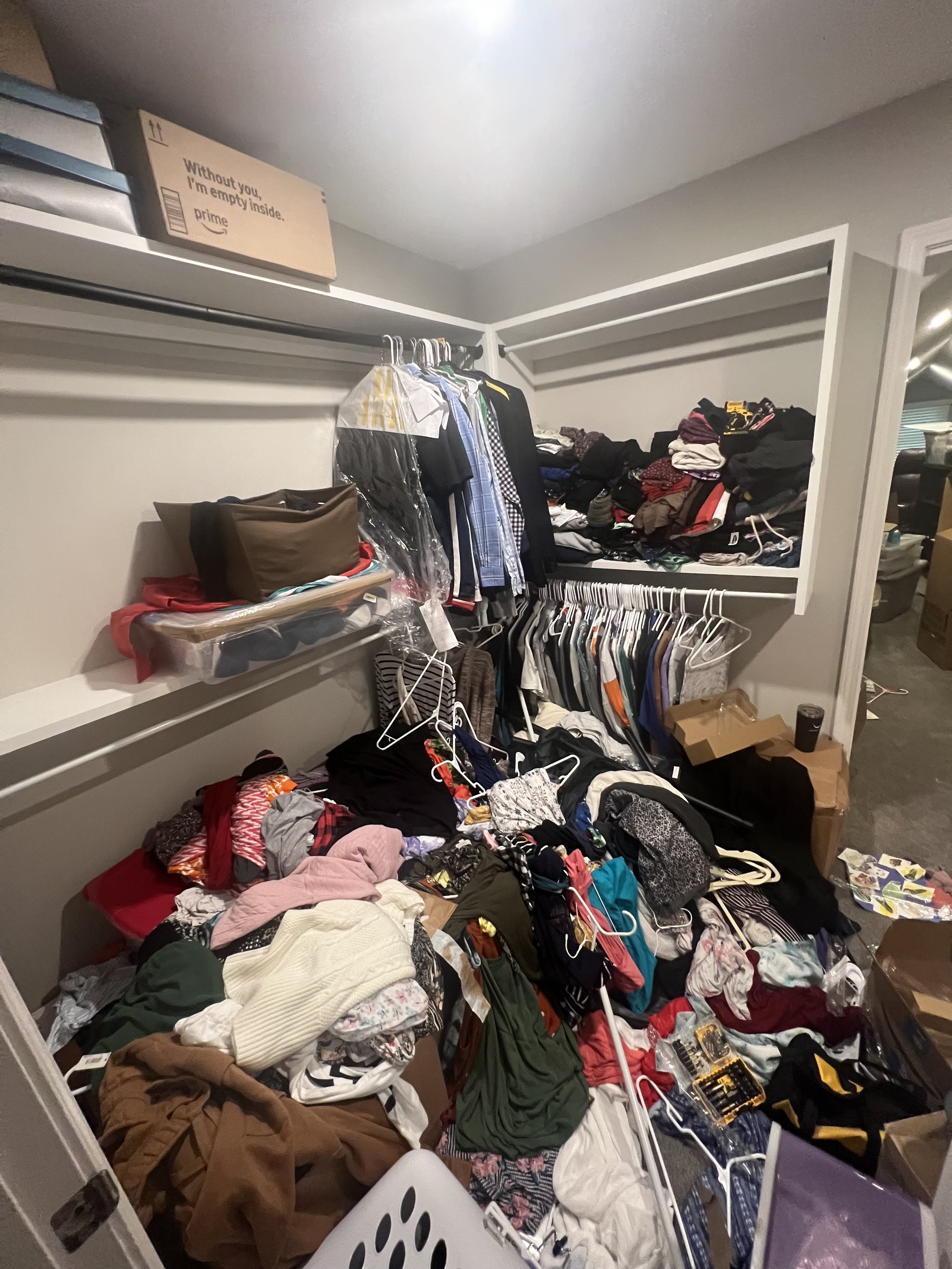 A messy walk-in closet with clothes and miscellaneous items scattered across the floor and hanging on rods, with boxes and piles of clothes on shelves and in the corner.