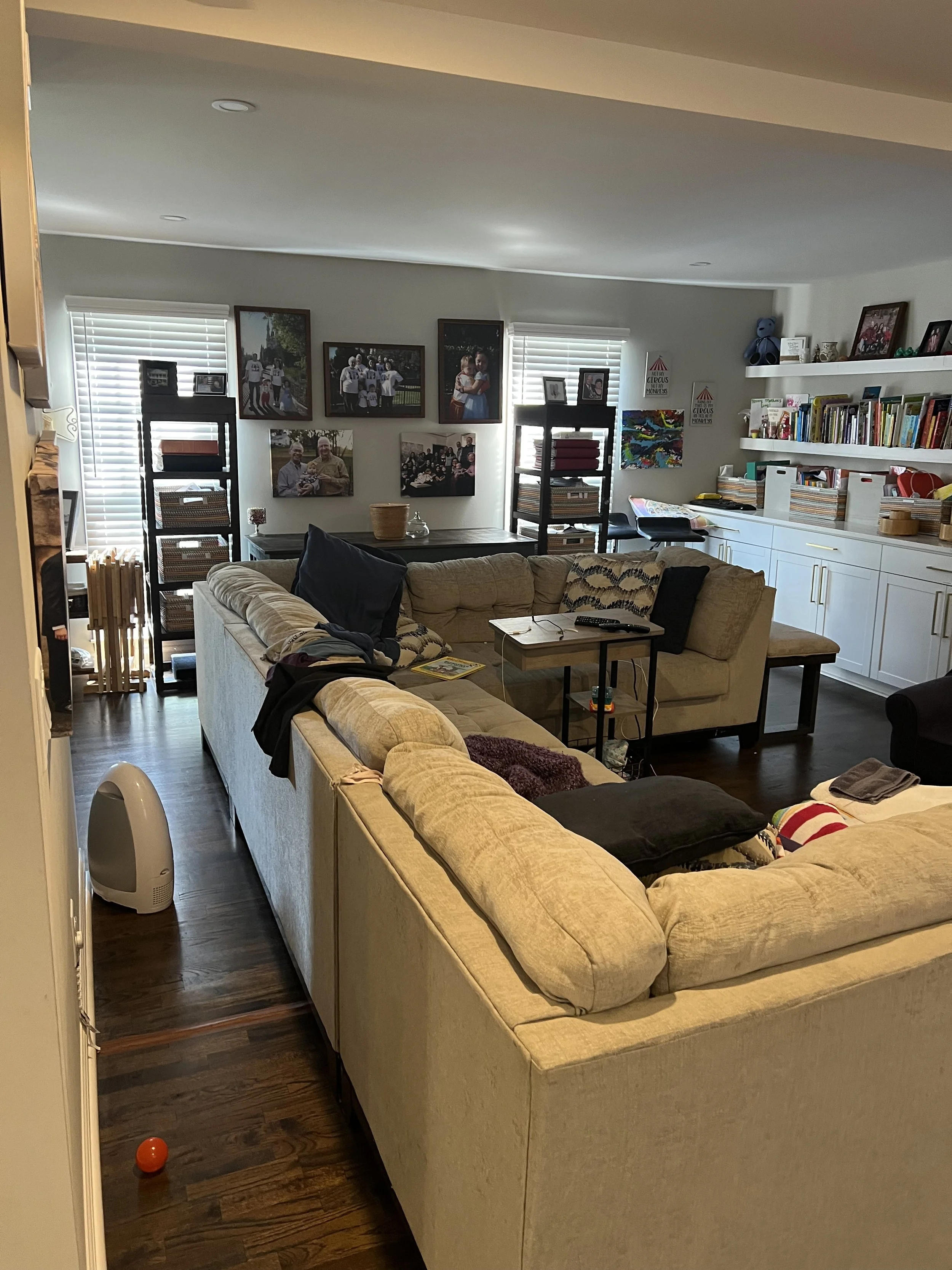 A living room with beige sectional sofa, TV remote, and black pillows, situated on a dark wooden floor. In the background are framed photographs on the wall, two black shelves with books and decorative items, and white cabinets with shelves holding b