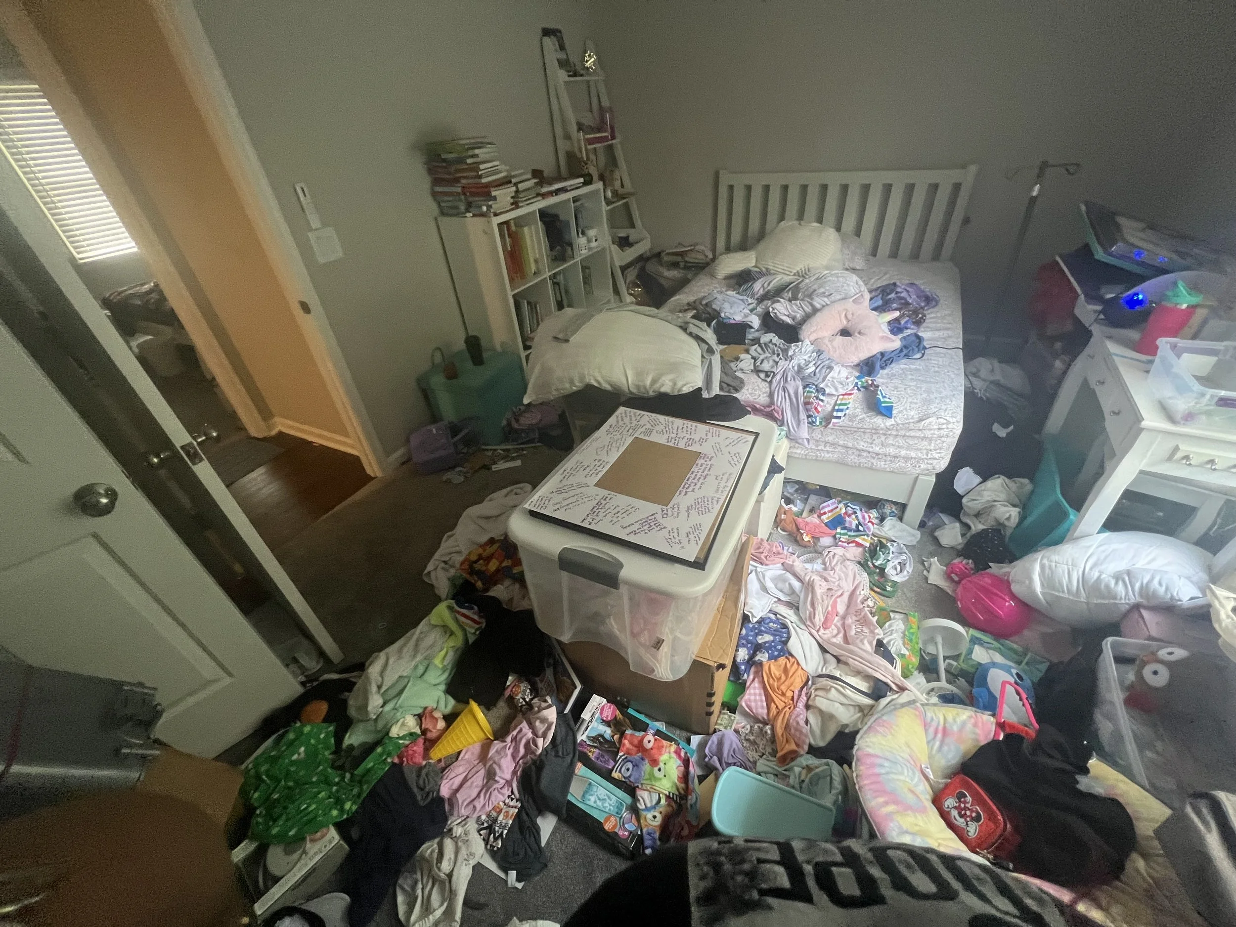 Disorganized bedroom with clothes, books, and belongings scattered on the floor, bed, and furniture.