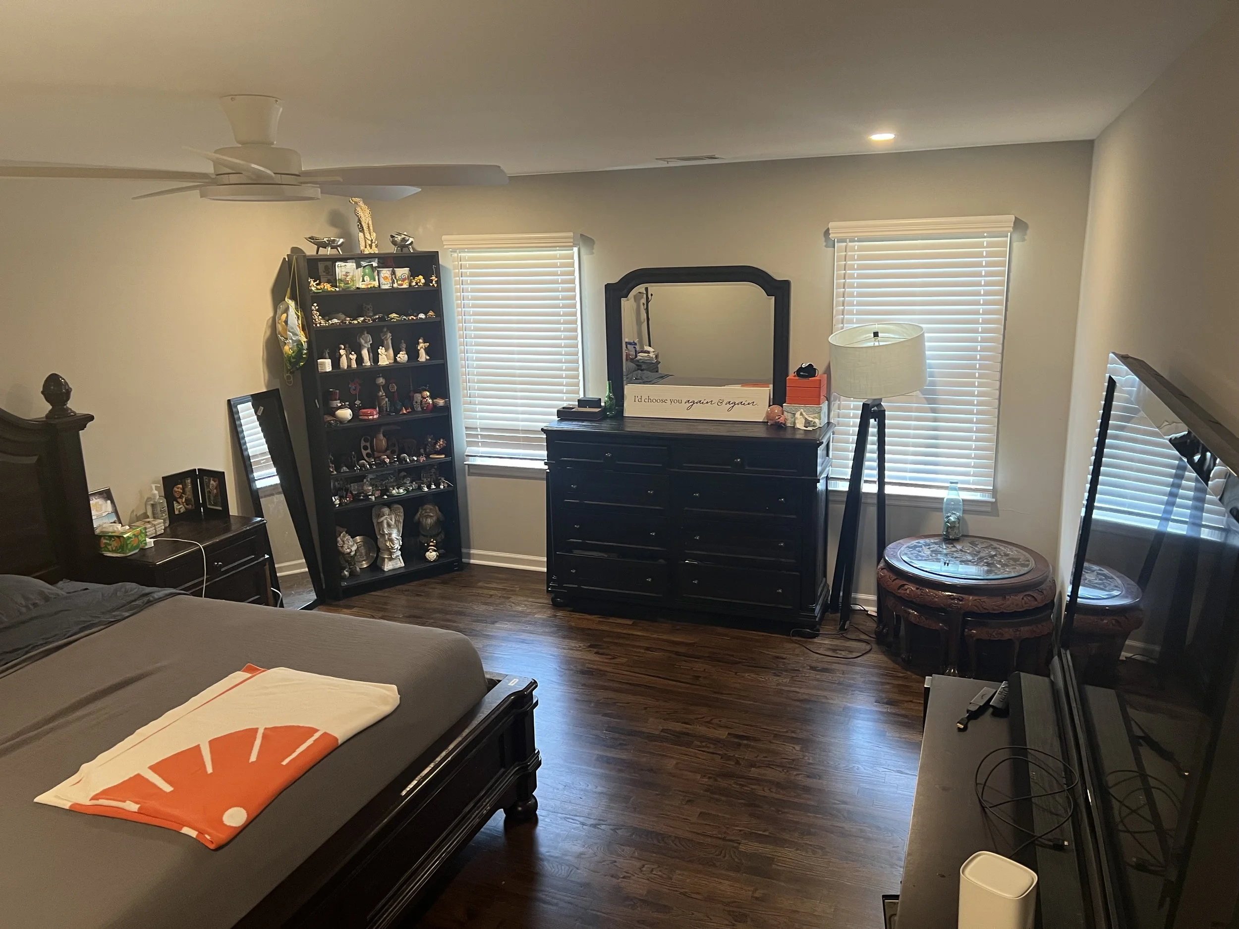 A bedroom with hardwood floors, a bed with a Towson University towel, a dark wooden nightstand with pictures and lotion, a tall black bookshelf with figurines and decorative items, a large black dresser with a mirror and a quote sign, two windows wit