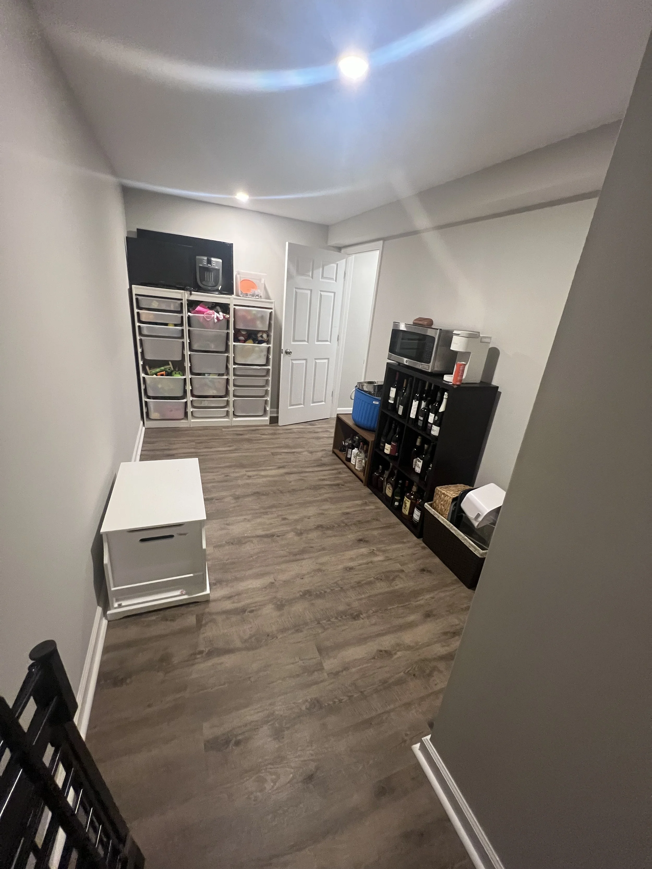 A small room with light-colored walls and wood floor, containing storage bins, a black wine rack with bottles, a microwave on top of the wine rack, a blue storage bin, and a white rolling cart.