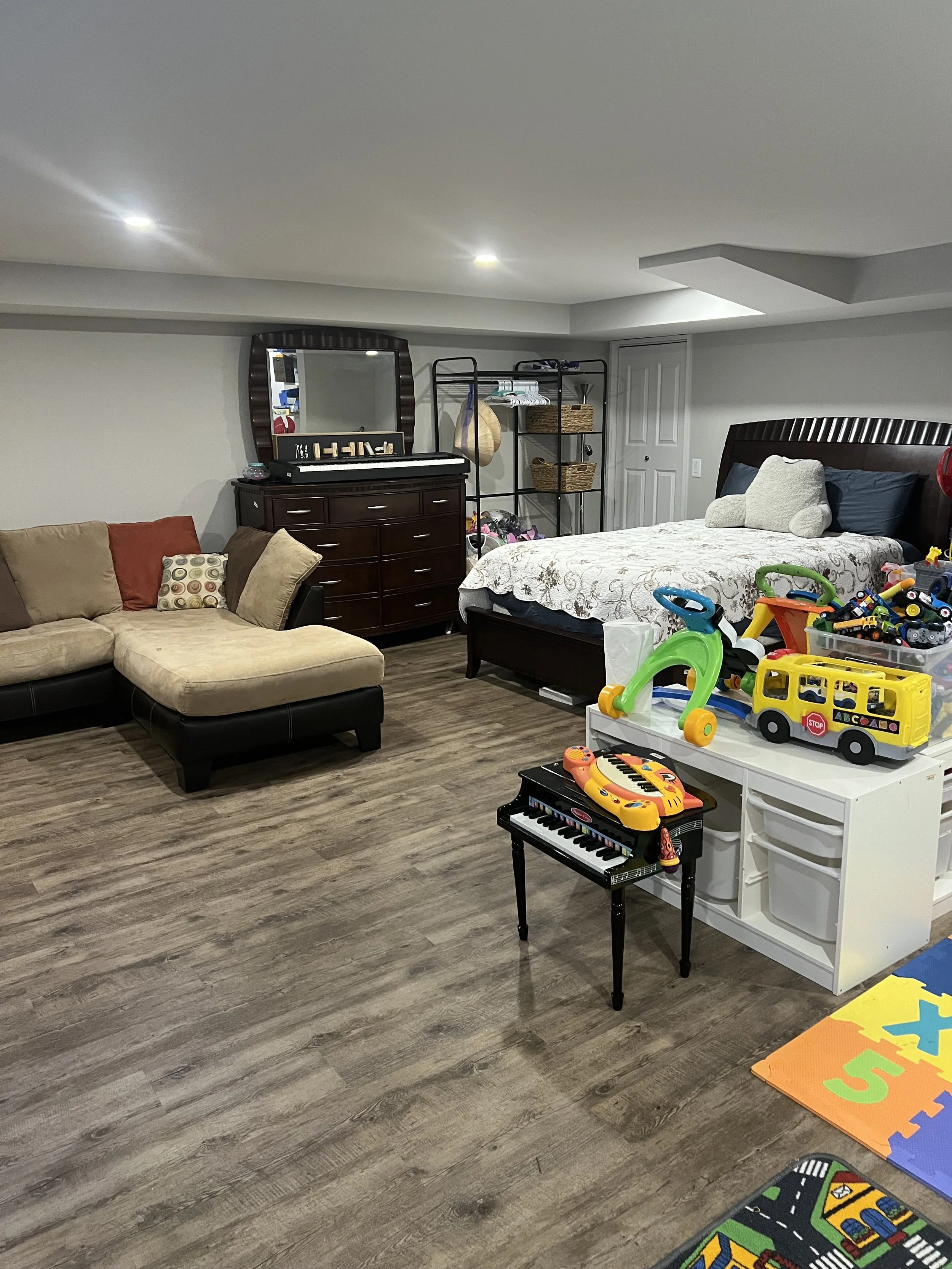 A basement room with a beige sectional couch on the left, a dark wooden dresser with a mirror behind it, a bed with a headboard to the right, and children's toys on a white storage unit and foam mats on the floor.