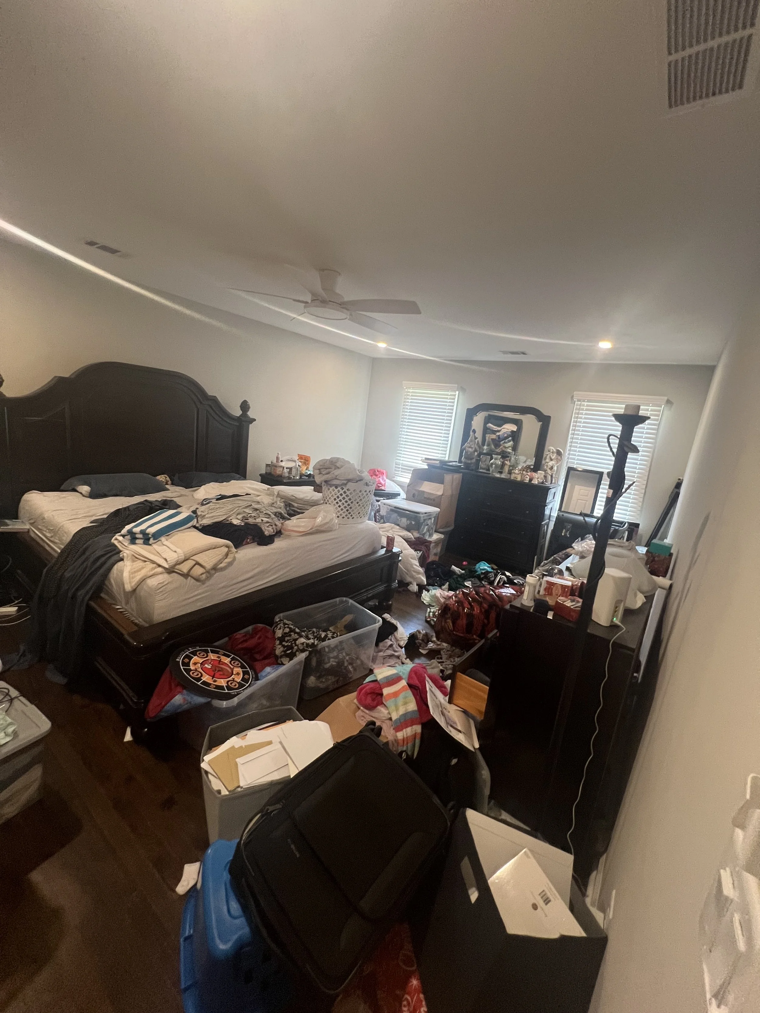 Cluttered bedroom with a large bed, multiple plastic bins, boxes, and various items scattered across the floor and furniture, including laundry, books, and decorative objects.