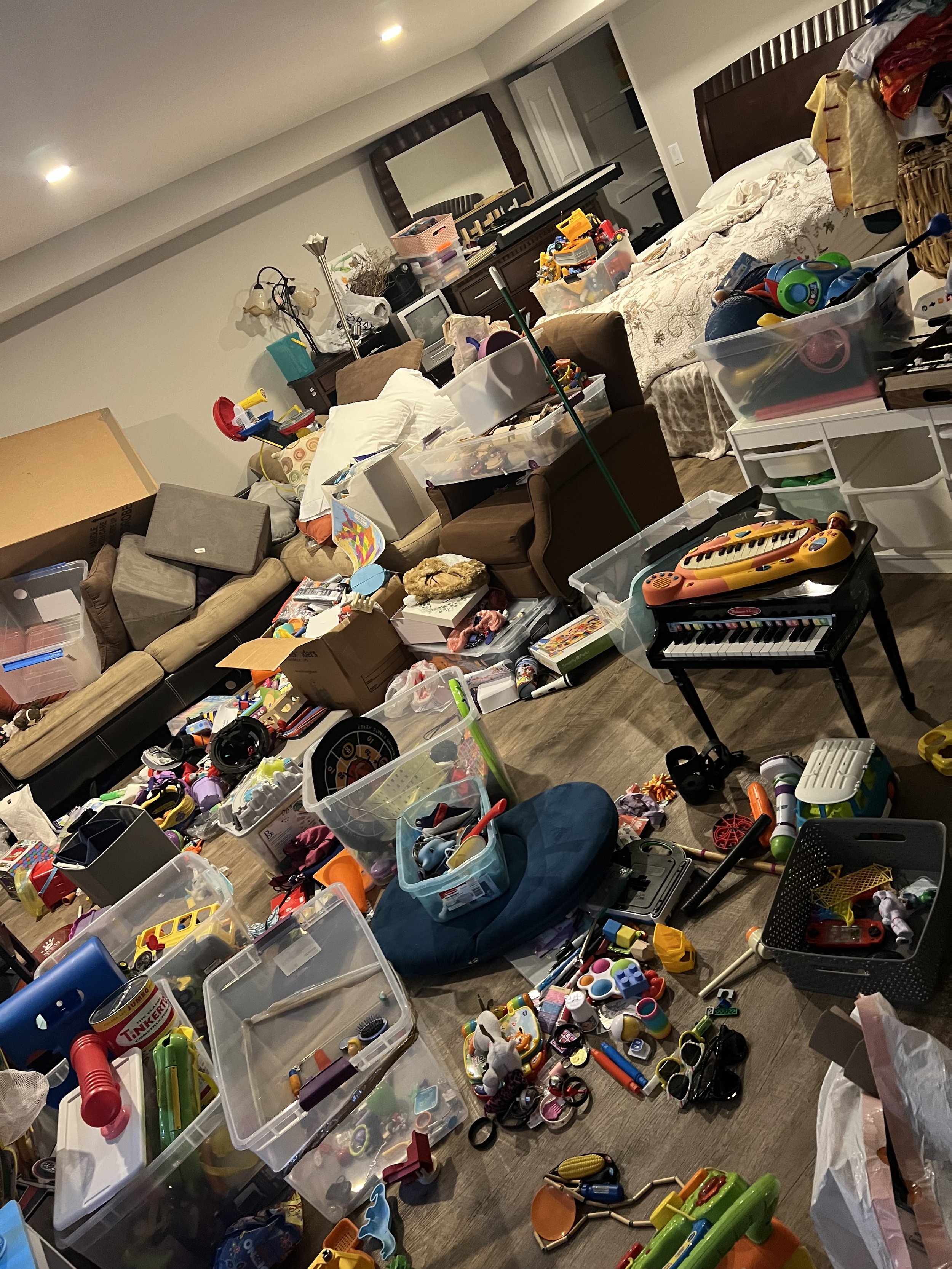 Living room with clutter and toys scattered across the floor, furniture, and a bed in the background.
