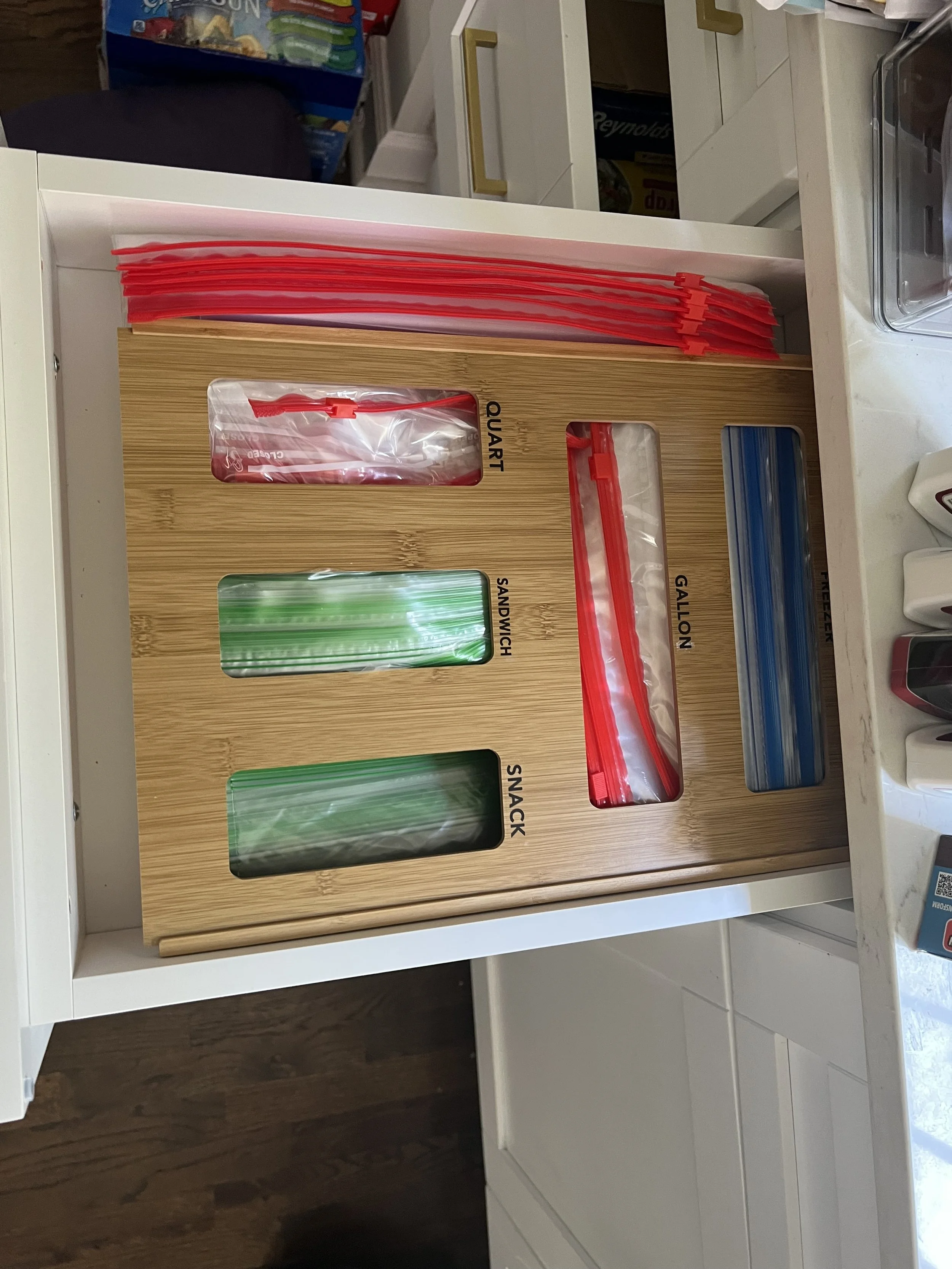Drawer organized with colorful plastic zipper bags labeled 'QUART,' 'SANDWICH,' 'SNACK,' 'GALLON,' and 'STORAGE' on a wooden organizer tray.