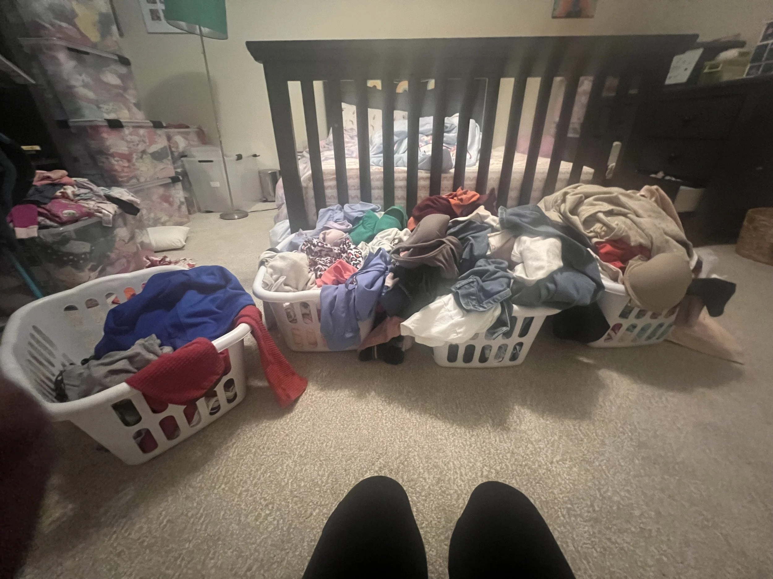 A room with a bed, laundry baskets filled with clothes, and various furniture and items in the background.