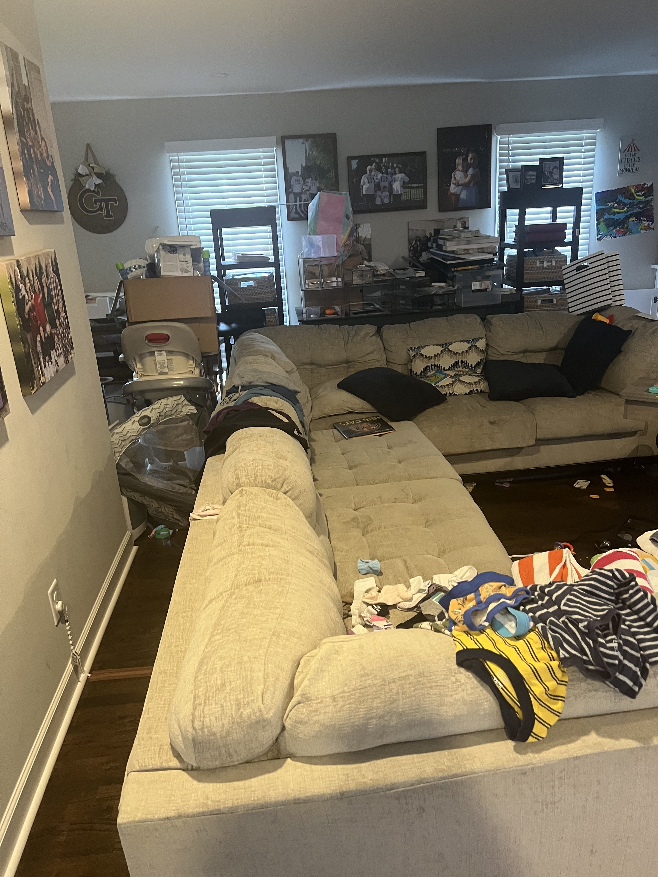 Living room with a beige sectional sofa covered with scattered clothes and accessories, black pillows, and a magazine. In the background, there are two windows with blinds, a wall adorned with framed photos, shelves with books and items, and a dining